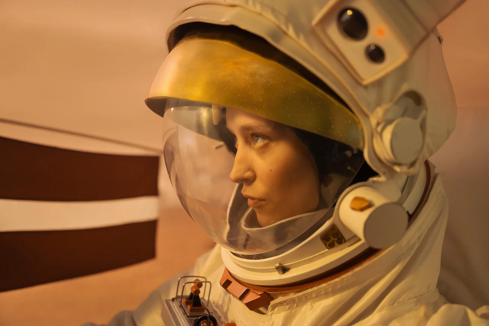 LMT reklāma — Close-up portrait of a woman in a white astronaut suit with a gold-tinted visor, seen from the side in warm, soft light. She appears contemplative inside a softly lit interior with muted brown and white striped shapes in the background;