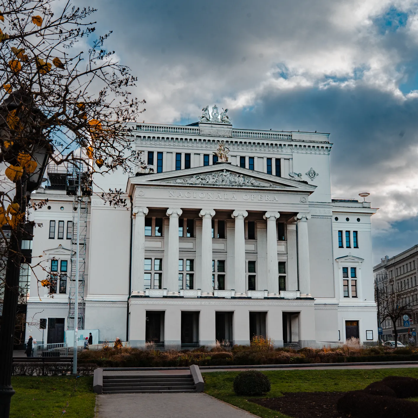 A white neoclassical opera building with a colonnaded portico and the inscription “NACIONALA OPERA” stands under a dramatic cloudy sky. In the foreground, a tree with yellow leaves, a lamppost, trimmed grass and steps lead toward the grand façade, captured in an architectural exterior style.