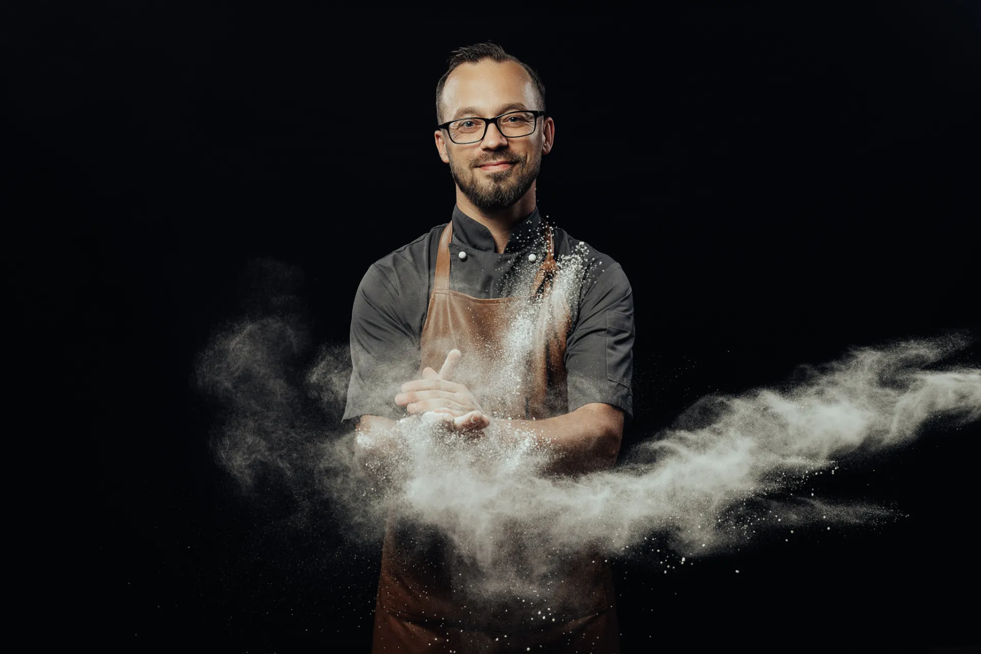 A smiling bearded man with glasses wearing a dark chef shirt and brown apron stands against a black studio background, clapping his hands as a cloud of white flour bursts outward. The high-contrast commercial portrait has a dramatic, dynamic mood.