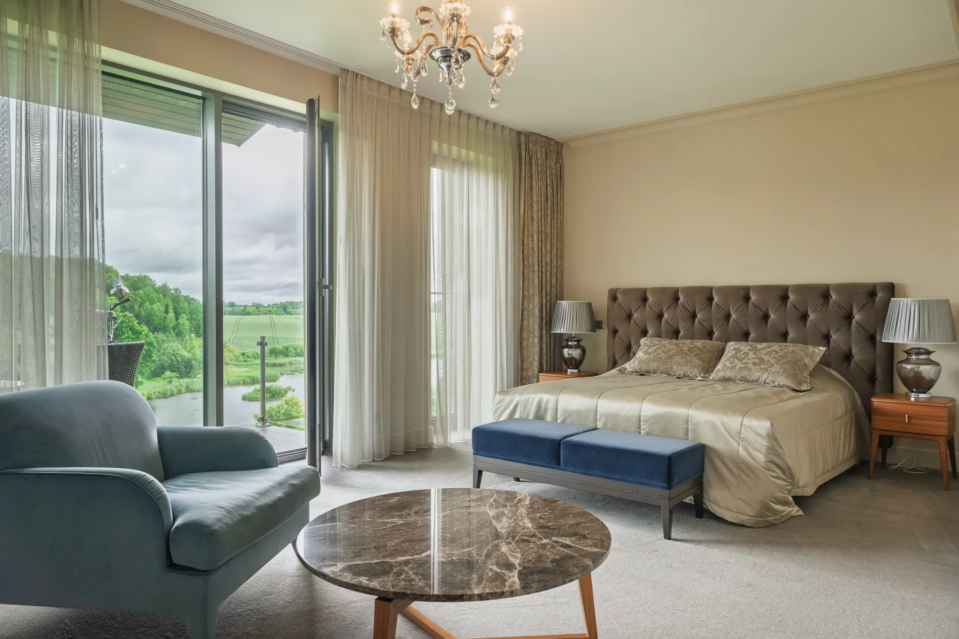 A bright, upscale bedroom interior with a large window view of fields and trees. A made bed with tufted headboard, blue bench, armchair, marble coffee table and crystal chandelier are visible under soft natural light. The style is a commercial interior/real estate photograph with a calm, luxurious mood.