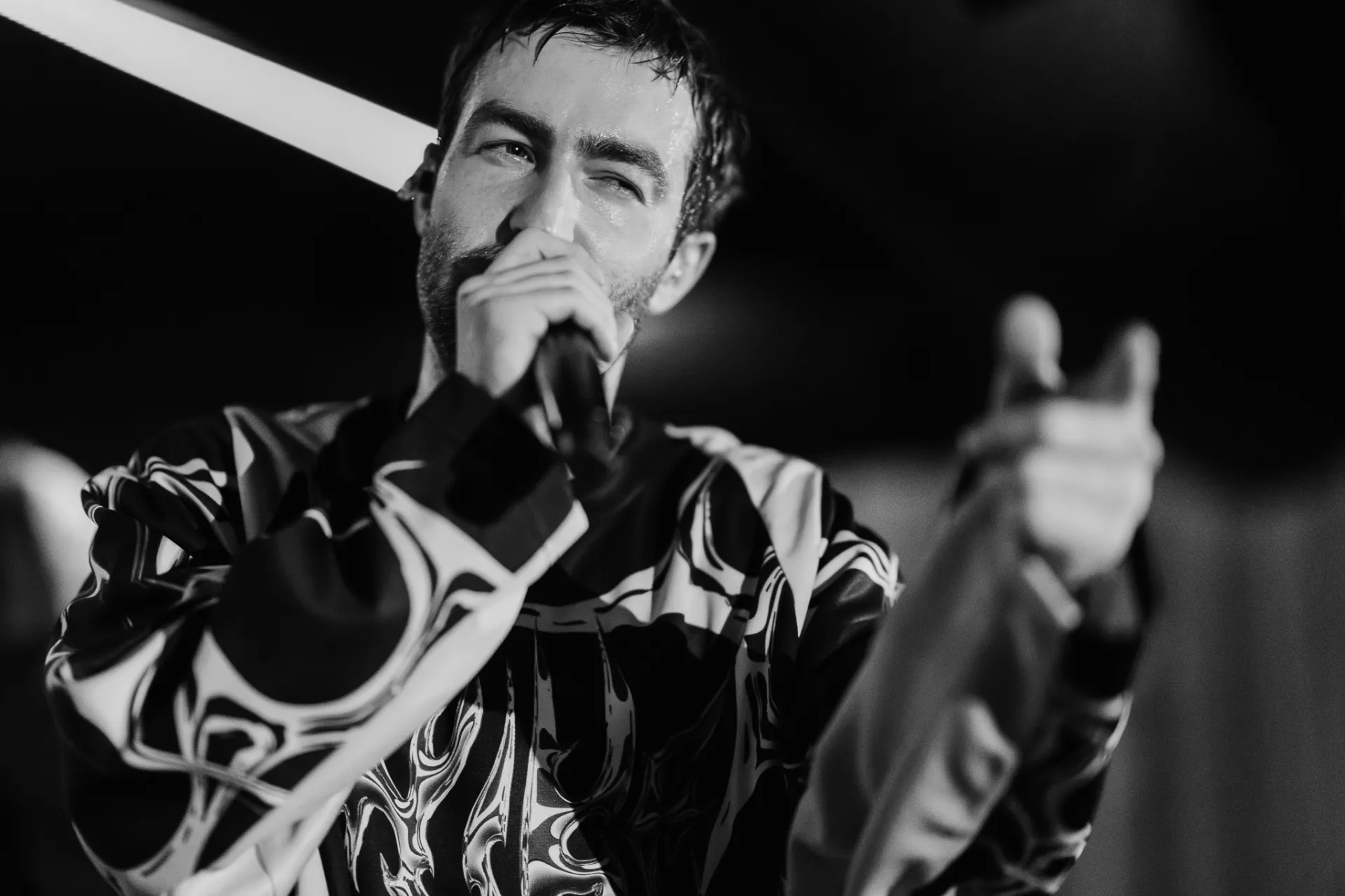 A black-and-white portrait of a bearded man singing into a microphone on stage, wearing a patterned shirt and gesturing with one hand under dramatic lighting.