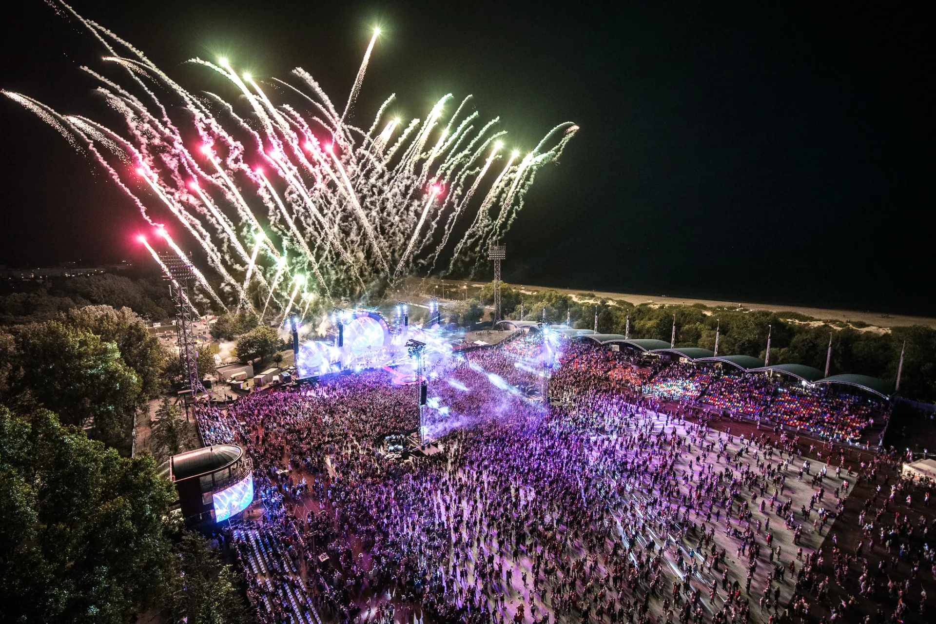 An aerial night shot of a large outdoor music festival with a dense crowd facing a brightly lit stage, while colorful fireworks burst in the dark sky above.