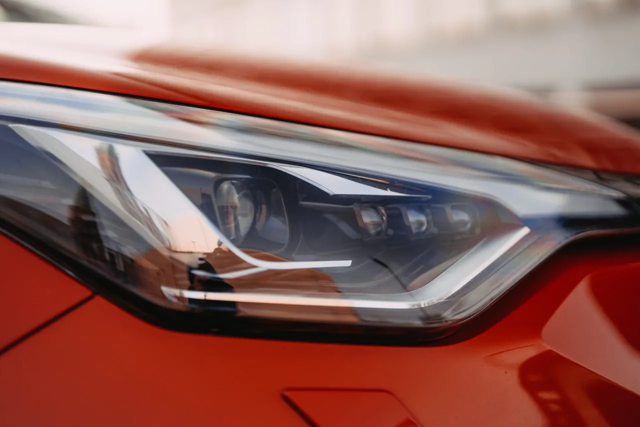 Auto Foto — Close-up of a red car headlight with sleek LED daytime running lights and chrome trim, captured in soft natural light with a shallow depth of field.