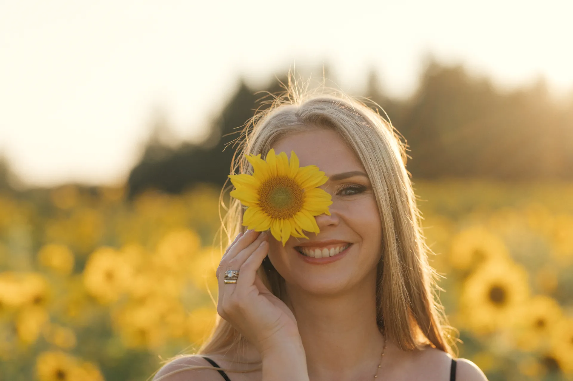 A smiling woman holds a sunflower in front of her face in a sunlit field of yellow flowers, with soft golden backlighting creating a warm portrait.