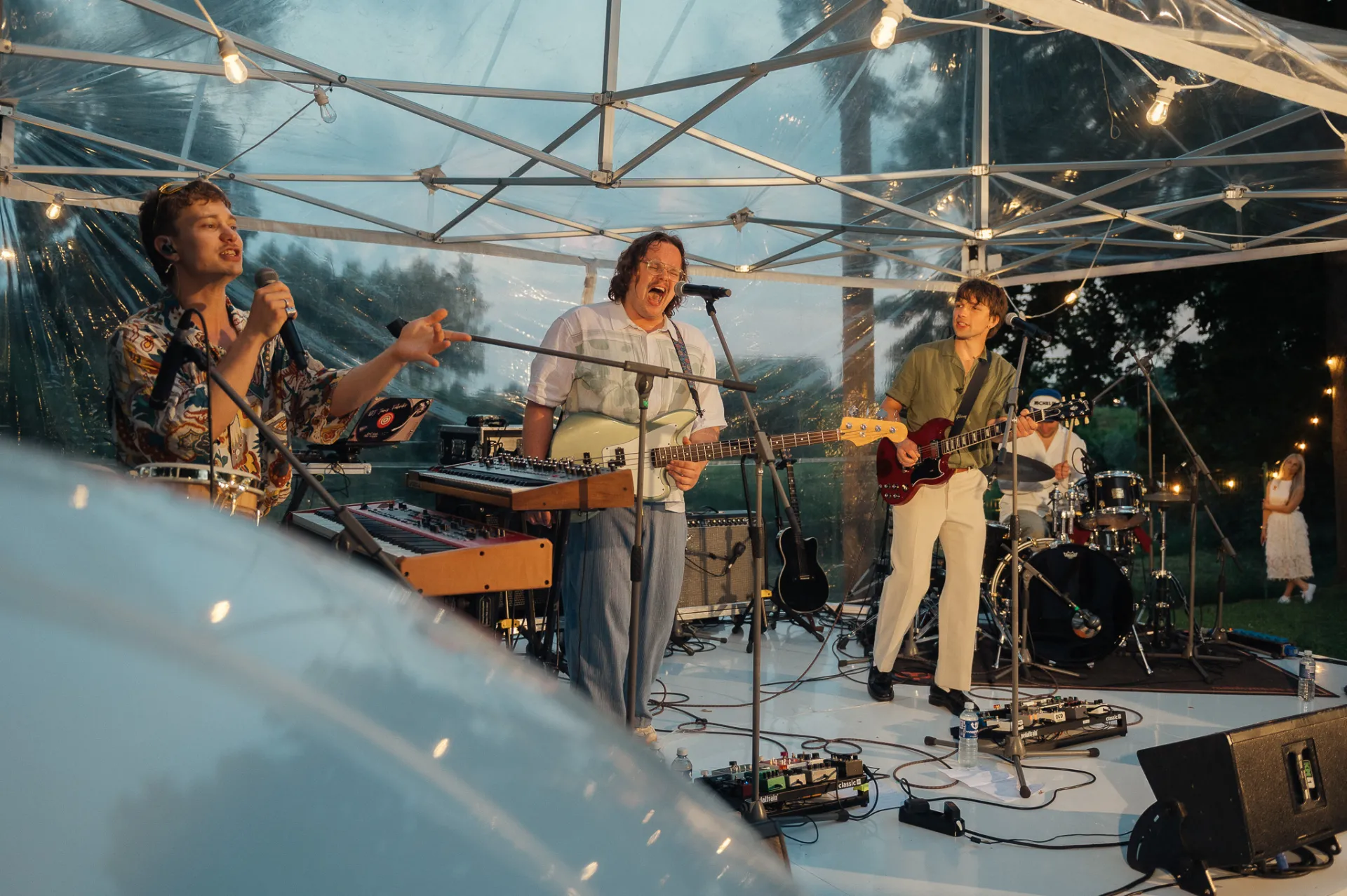 Three musicians perform on a white stage under a clear tent with string lights at dusk. A singer with a microphone gestures while a bassist and guitarist play, with keyboards, drums and gear visible. The outdoor concert scene has a lively, warm evening mood, captured in a live music event style.