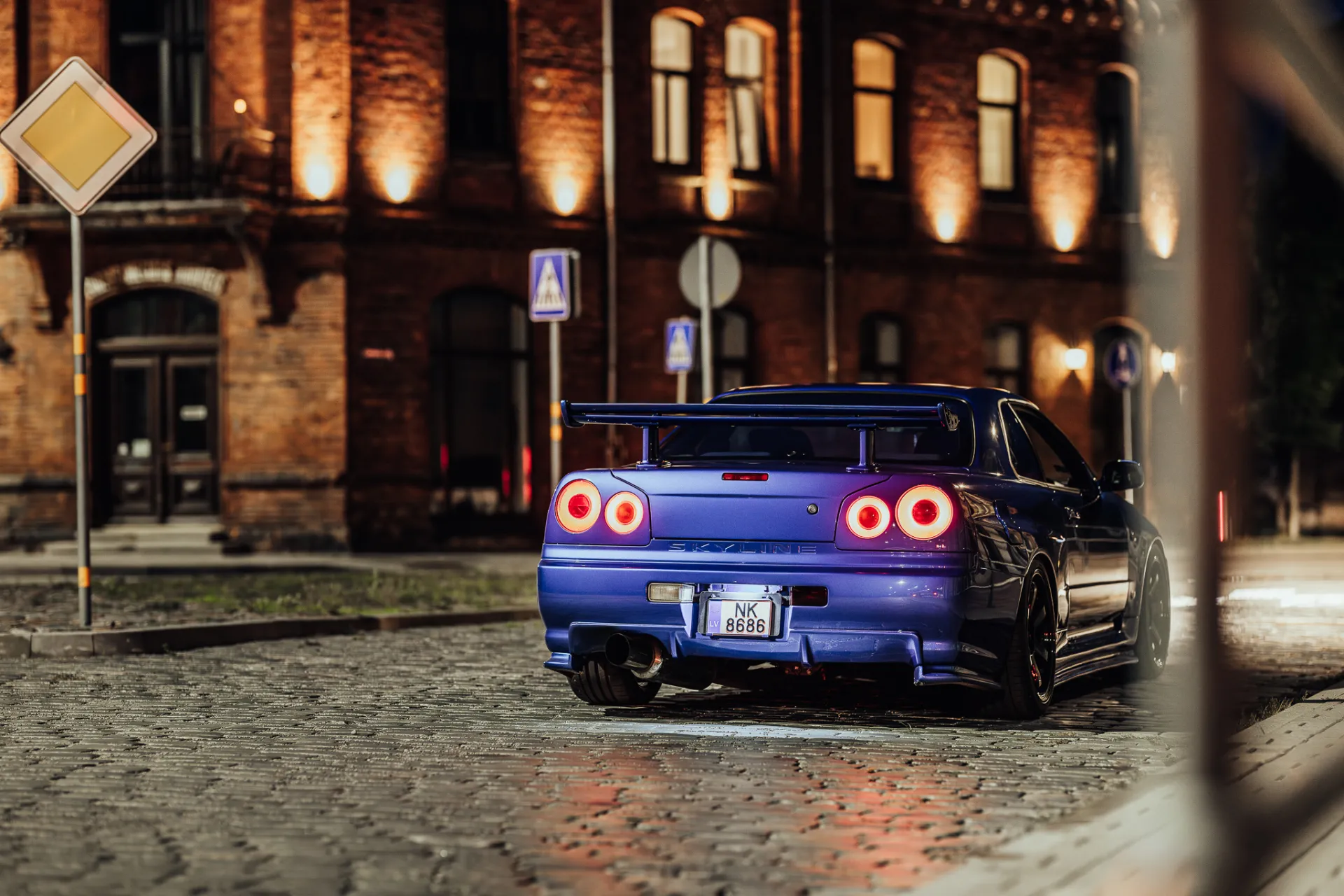 A blue sports coupe with a large rear wing and four illuminated circular taillights is parked on a cobblestone street at night, viewed from the rear three-quarter angle. Warm uplighting highlights a brick building with arched windows and doors behind it, creating a moody urban automotive night scene.