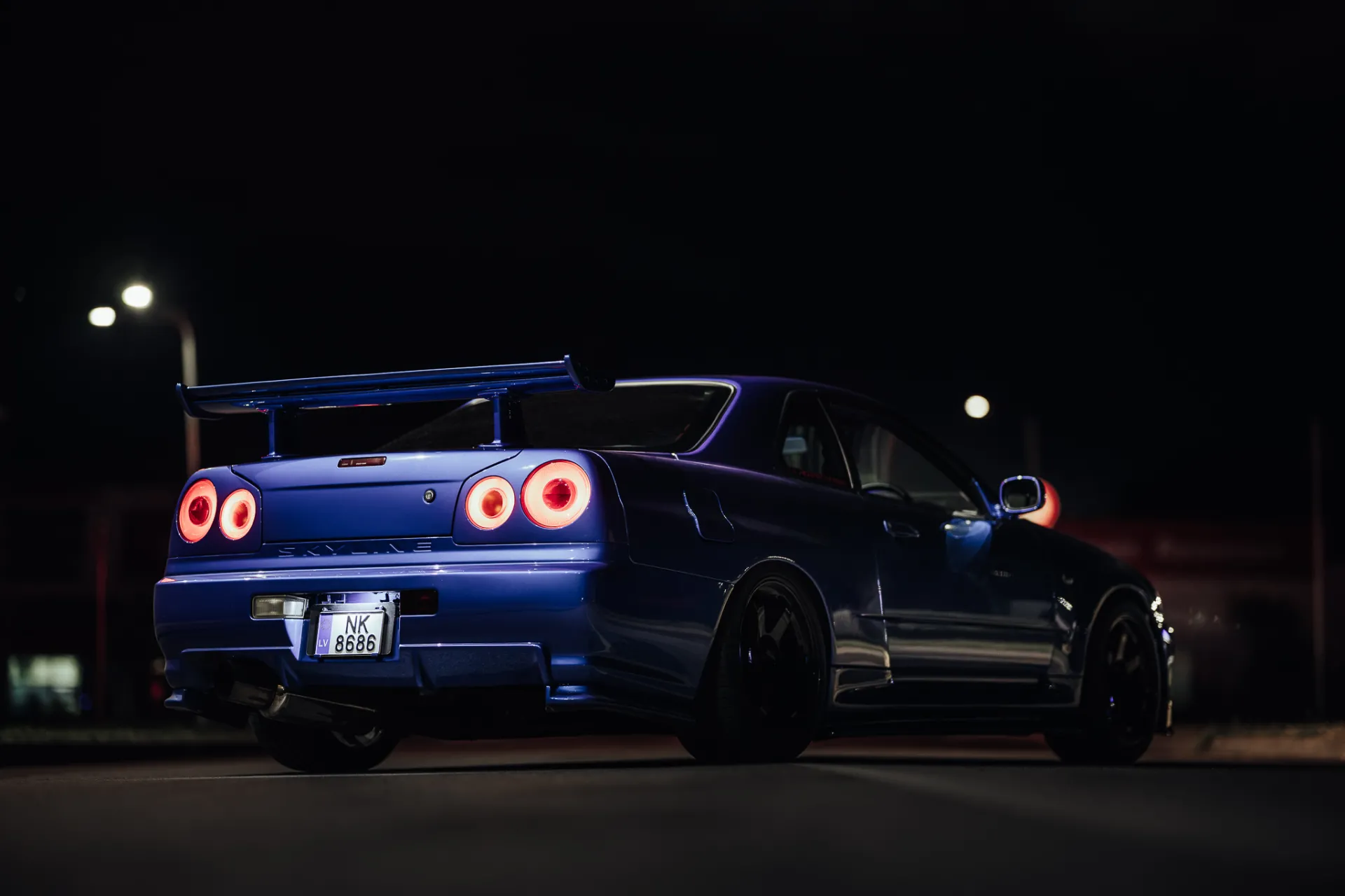 A blue sports car with a large rear wing is shown from behind at night, its circular taillights glowing red against the dark asphalt. Streetlights blur in the background, creating a moody automotive portrait.