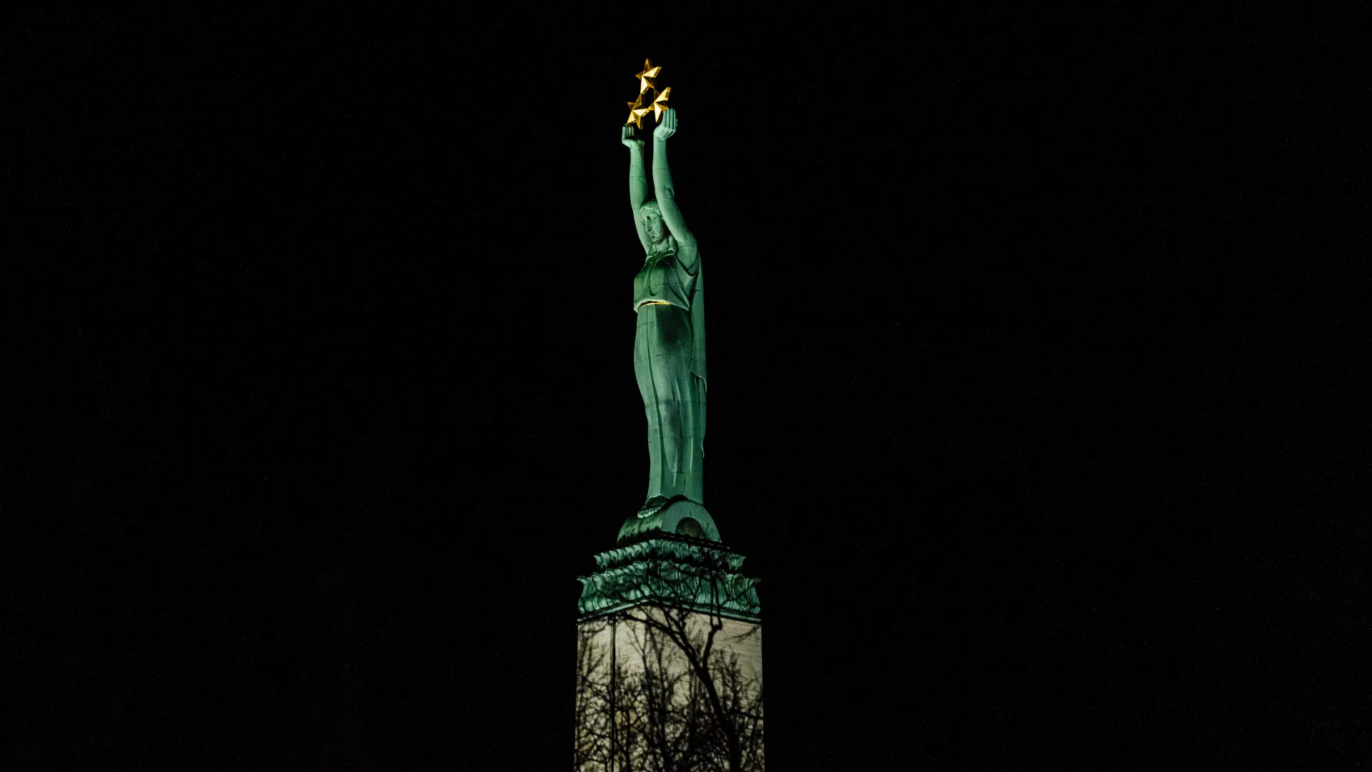 Naktī uz akmens pjedestāla stāv zaļa statuja, kas ar paceltām rokām tur zelta zvaigznes pret tumšo debesi, bet zem tās izceļas kailu koku zaru silueti.