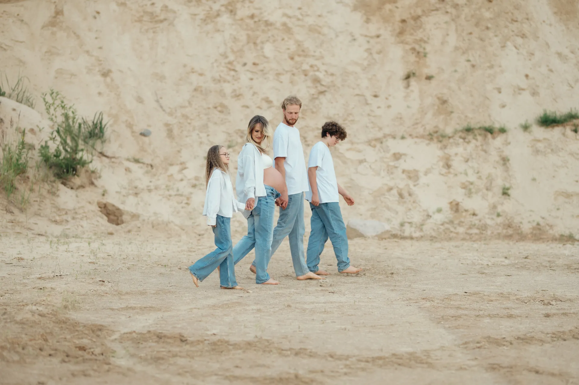 A pregnant woman walks barefoot in line with a man and two children on sandy ground in front of a pale, eroded cliff. All wear white tops and light blue jeans, moving left to right in soft natural light. The lifestyle family portrait has a calm, warm mood.