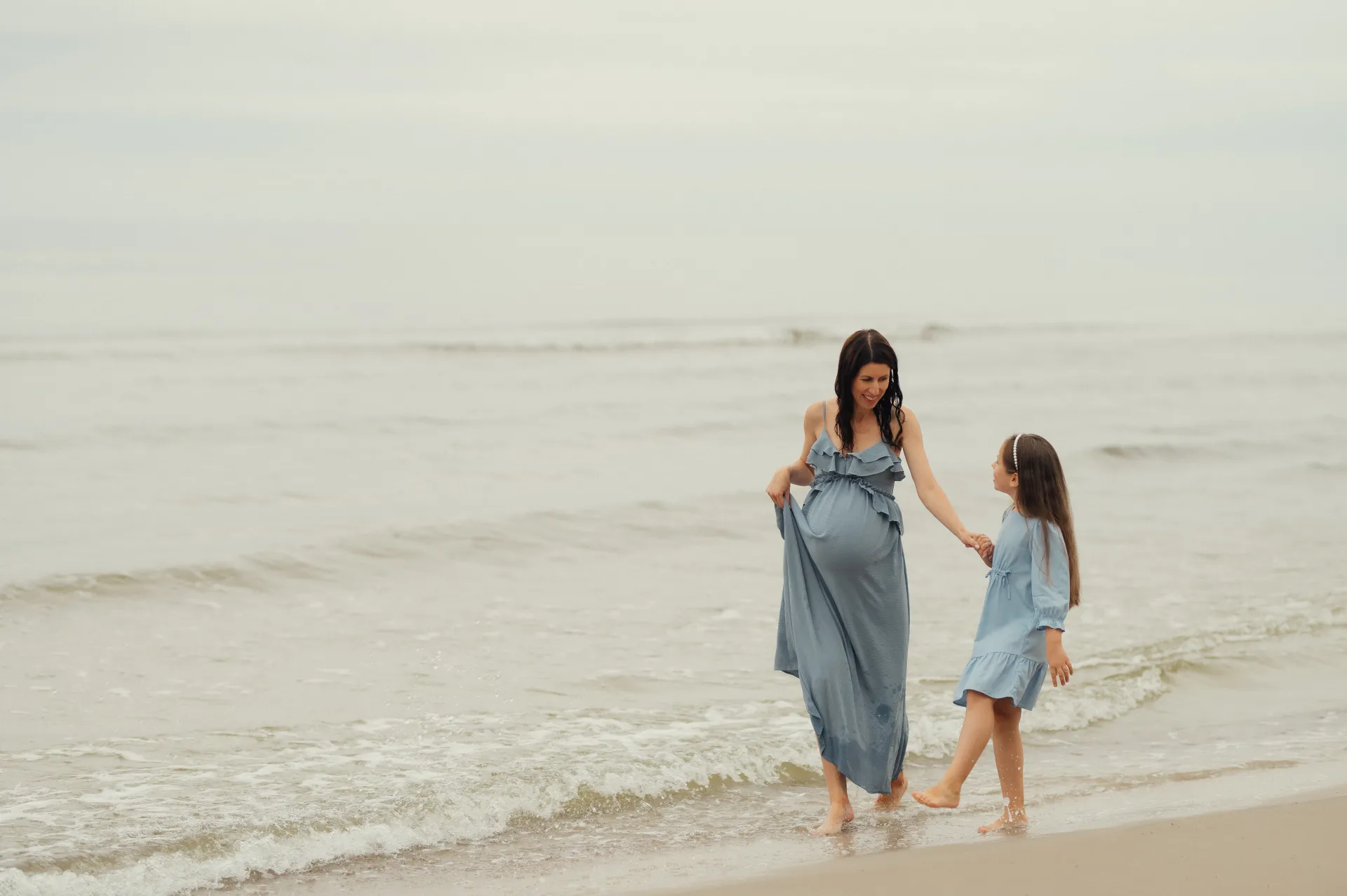 A pregnant woman in a long blue ruffled dress holds the hand of a young girl in a light blue dress as they walk barefoot along a sandy shore with gentle waves. The overcast seaside setting and soft light create a calm, intimate maternity portrait mood.