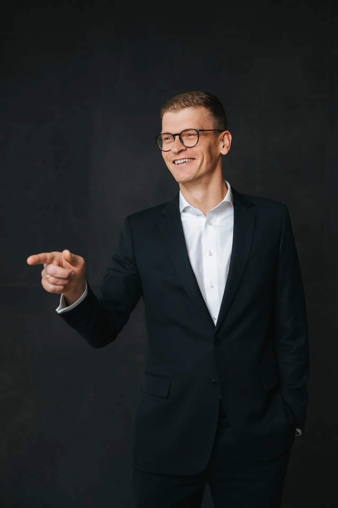 Biznesa Portreti — Smaidošs vīrietis ar brillēm un īsiem gaiši brūniem matiem melnā uzvalkā, zem kura redzama balta krekla ar pogām, stāv uz tumša, faktūriem bagāta fona. Viņš stāv studijas portreta pozā, ar labo roku rādot uz priekšu, bet kreiso roku tur kabatā; maigais studijas apgaismojums rada …