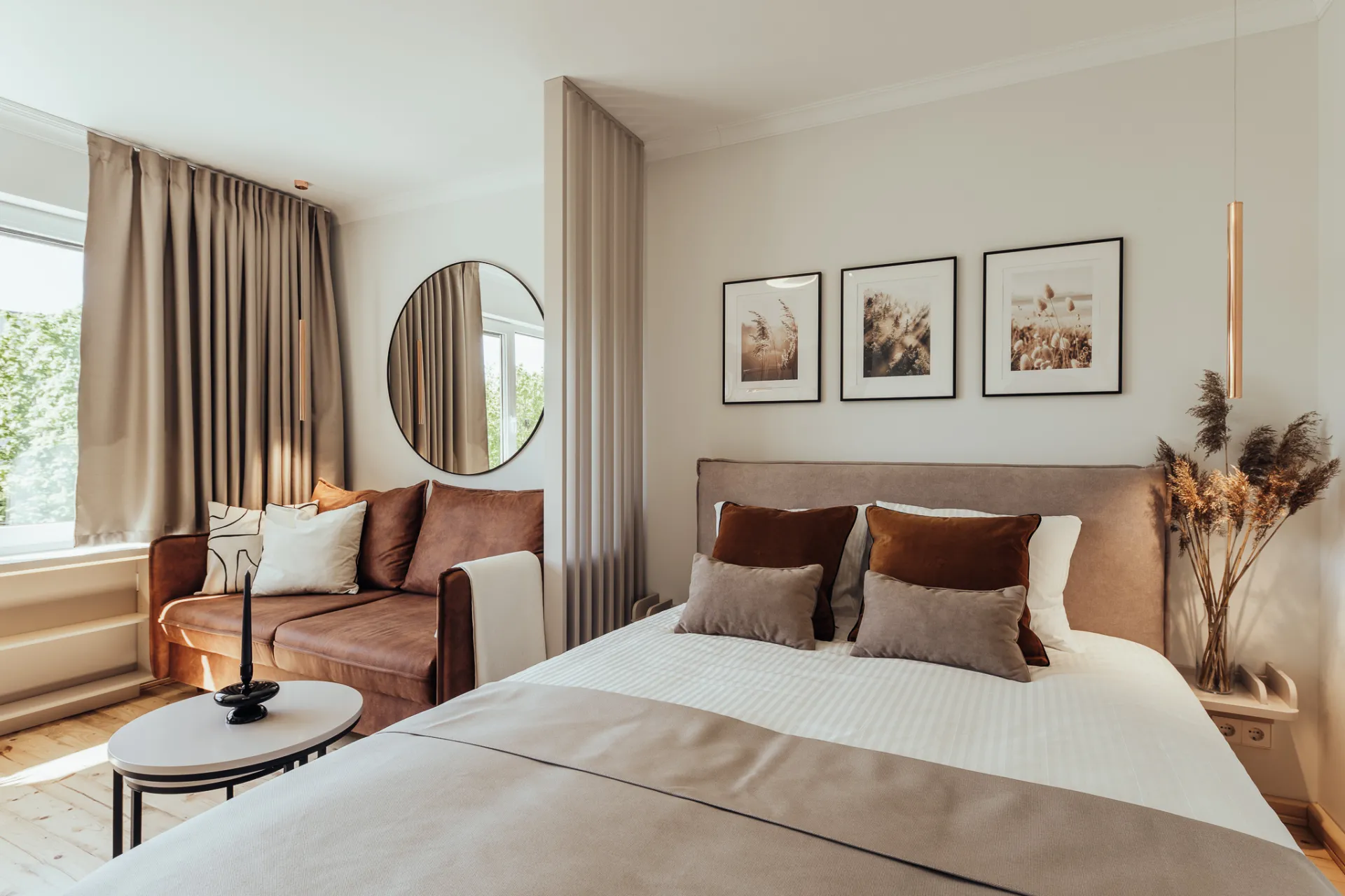 A bright, minimalist bedroom interior with a made bed featuring brown and taupe pillows and a neutral throw, framed botanical prints above the headboard and dried pampas grass on a nightstand. An adjacent sitting area with a brown velvet loveseat, round mirror and small side table by a curtained window.