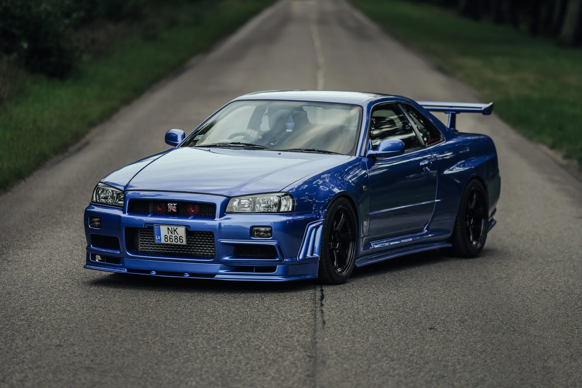A vibrant blue sports coupe with a large rear wing and black wheels is parked at a three-quarter front angle on a narrow asphalt country road flanked by grass and trees.