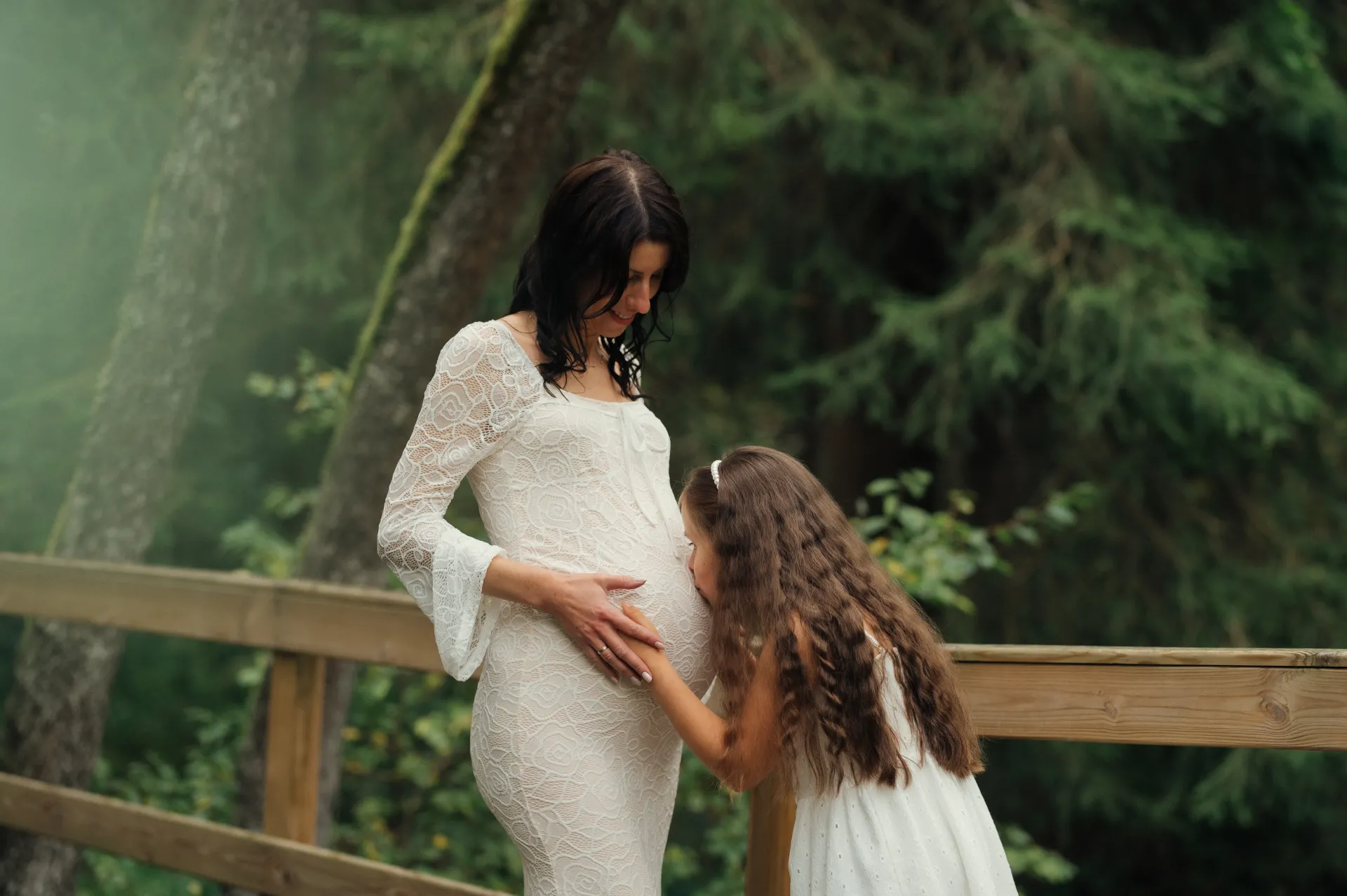 A pregnant woman in a white lace maternity dress stands outdoors beside a young girl in a white dress who leans in to kiss the woman’s belly. They are on a wooden railing in a misty green forest with soft natural light, creating a tender maternity portrait with a warm, intimate mood.