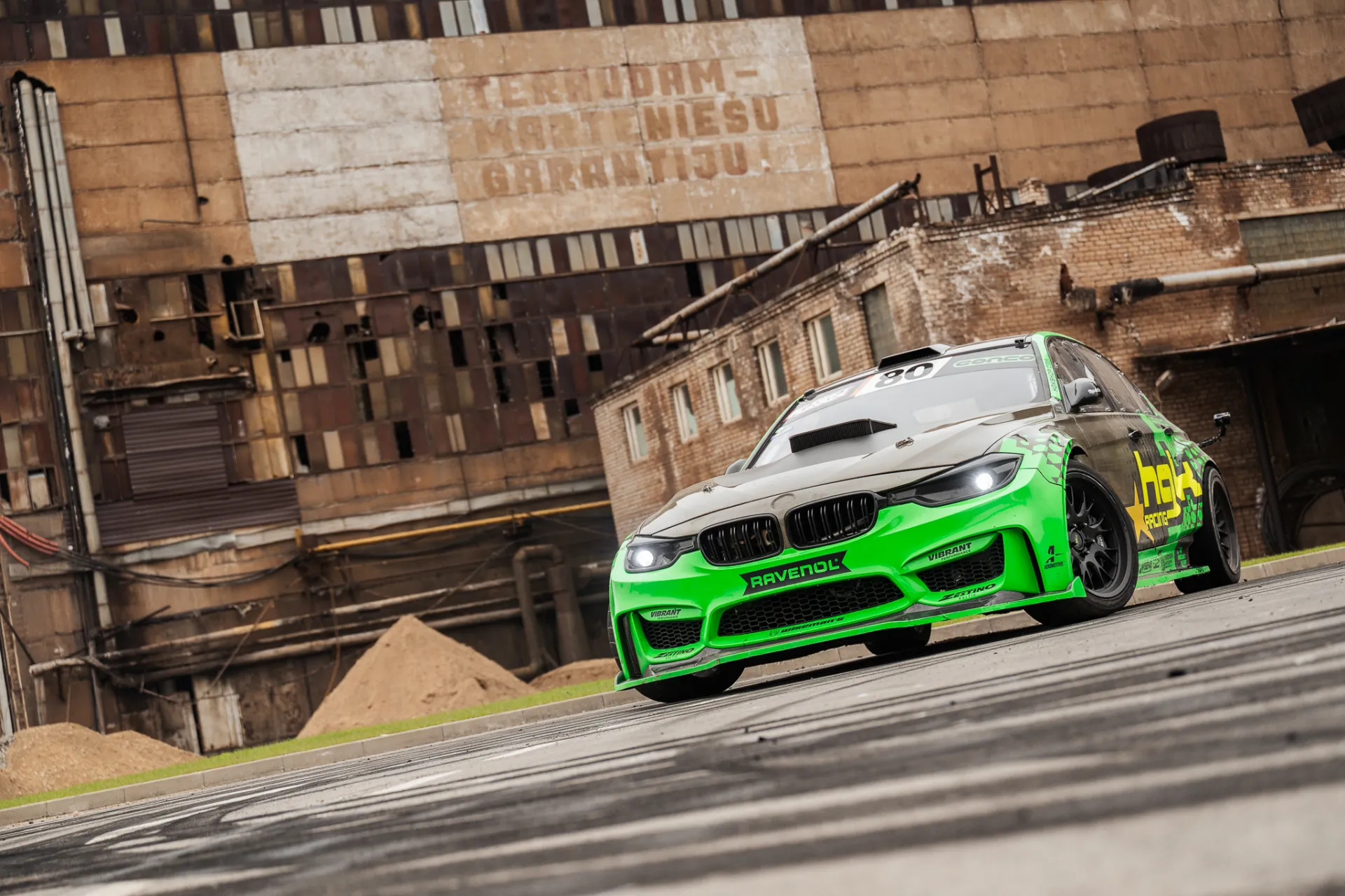Spilgti zaļš un melns BMW sacīkšu automobilis ar „Ravenol“ sponsoru uzlīmēm traucas pa asfalta trasi ar ieslēgtiem lukturiem, fonā redzama laika apstākļu ietekmēta rūpnieciskā ķieģeļu ēka ar izbalējušiem uzrakstiem apmākušā dienasgaismā.