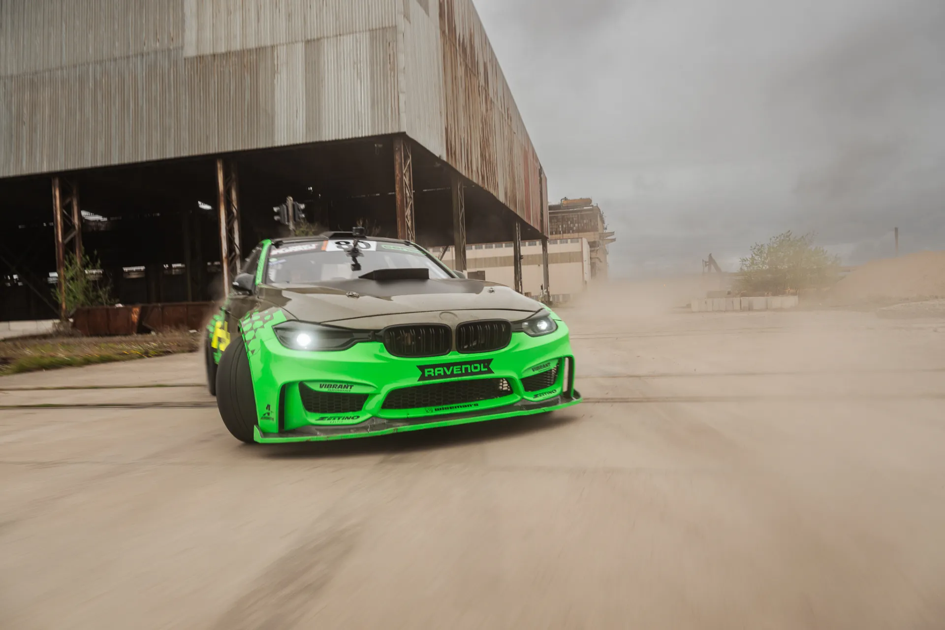 A bright green wide-body race car with a dark hood drifts forward on a dusty concrete lot, kicking up a plume of dust. It is framed in front of a large weathered industrial building under an overcast sky, captured with motion blur in a dynamic automotive commercial style.