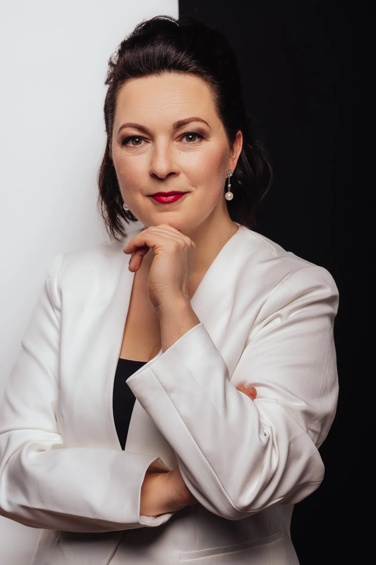 A woman with dark hair styled up wears a white blazer over a black top, resting her chin on her hand against a split white and black background. Soft studio lighting highlights her red lipstick and pearl earrings in a professional portrait.