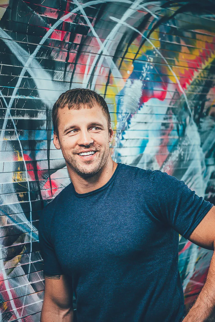 Mairis Briedis — A smiling man with short brown hair and light stubble wears a dark blue T-shirt, leaning slightly against a colorful graffiti-covered wall. The close-up portrait is lit with soft directional light, creating a casual, upbeat mood.