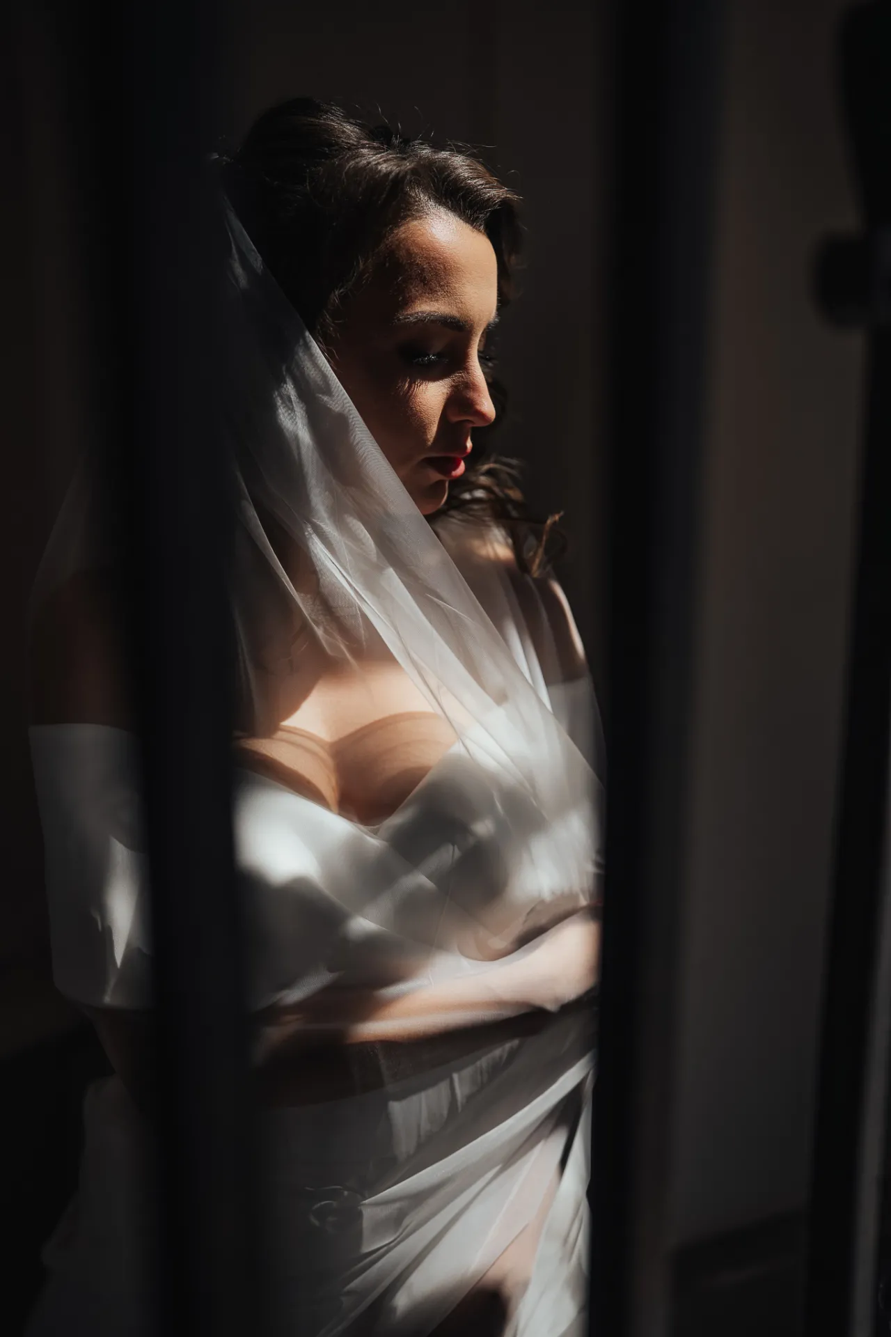 A bride in a white wedding gown and veil stands in profile in a dim interior, her eyes cast downward. Strong directional sunlight highlights her face and dress through dark vertical framing, creating a moody, dramatic wedding portrait with soft, cinematic contrast.