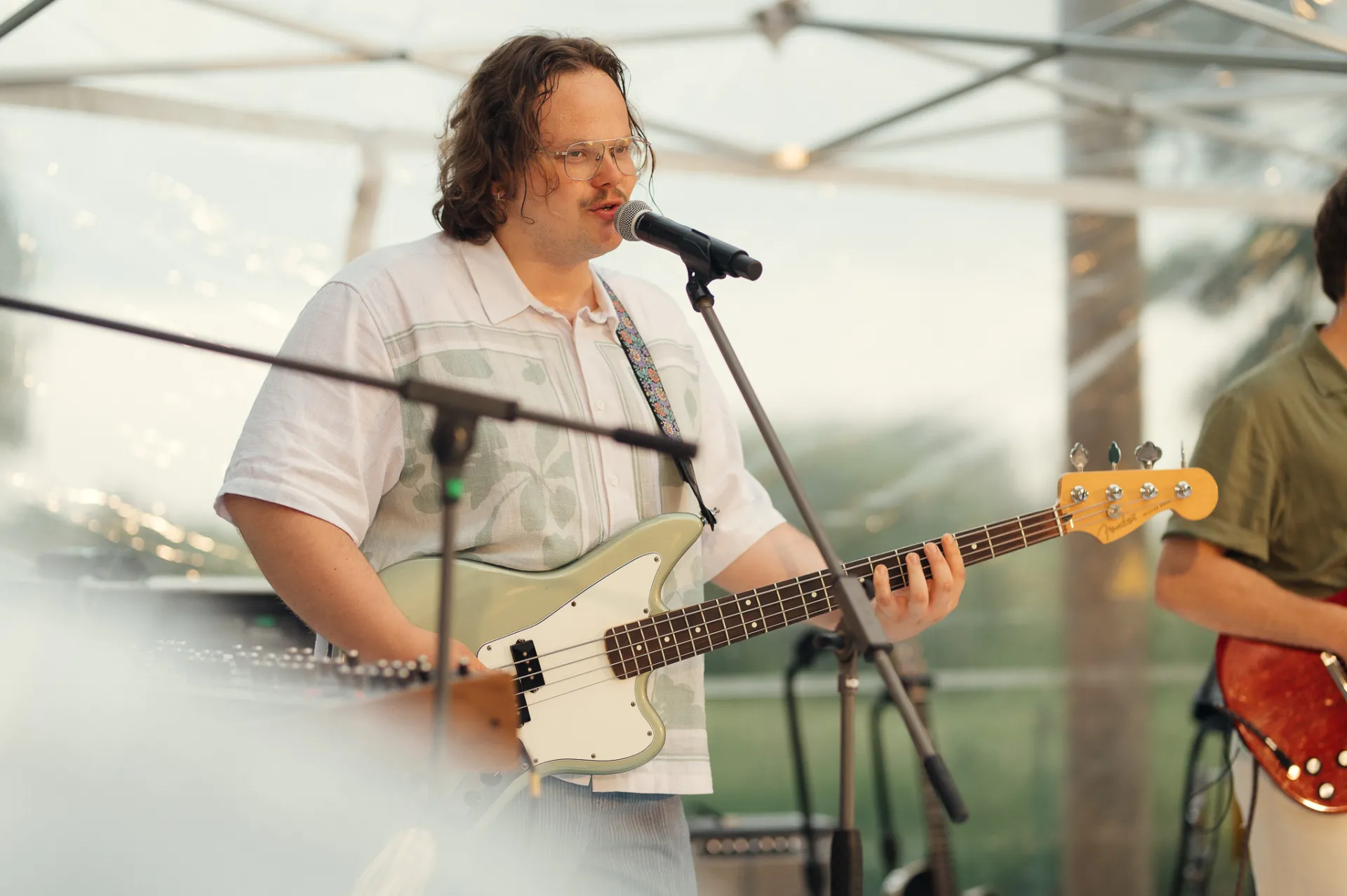 A musician with curly hair and glasses sings into a microphone while playing a light green bass guitar outdoors under a translucent canopy. A second guitarist is partially visible beside him; soft diffused light and light haze create a relaxed live performance atmosphere in a concert photography style.