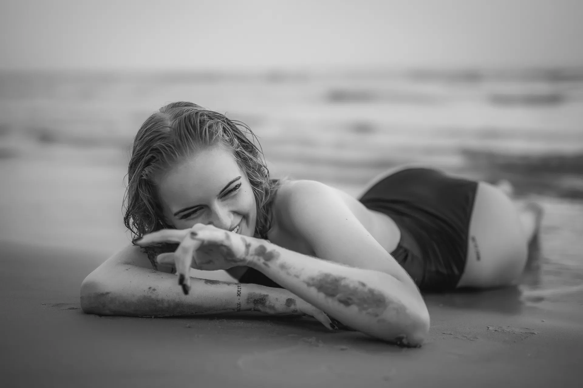A black-and-white portrait of a woman lying on wet sand at the beach, smiling with her head resting on her arms, wearing a dark swimsuit with ocean waves blurred in the background.