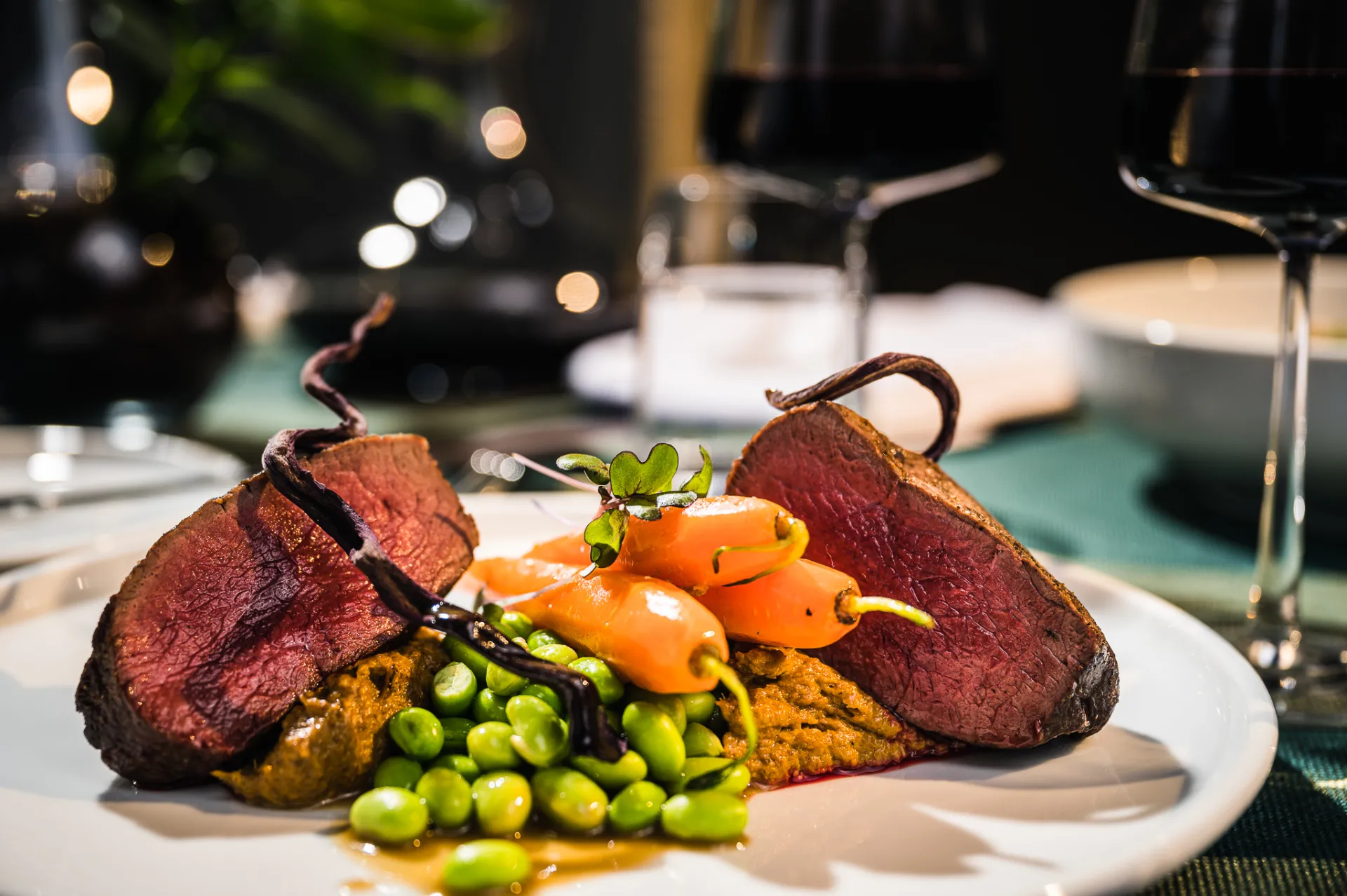 A close-up food photograph shows two slices of medium-rare roast meat on a white plate with baby carrots, edamame and a purée, garnished with a twisted vegetable and microgreens. The upscale dining setting with wine glasses and soft warm lighting conveys a refined commercial culinary style.