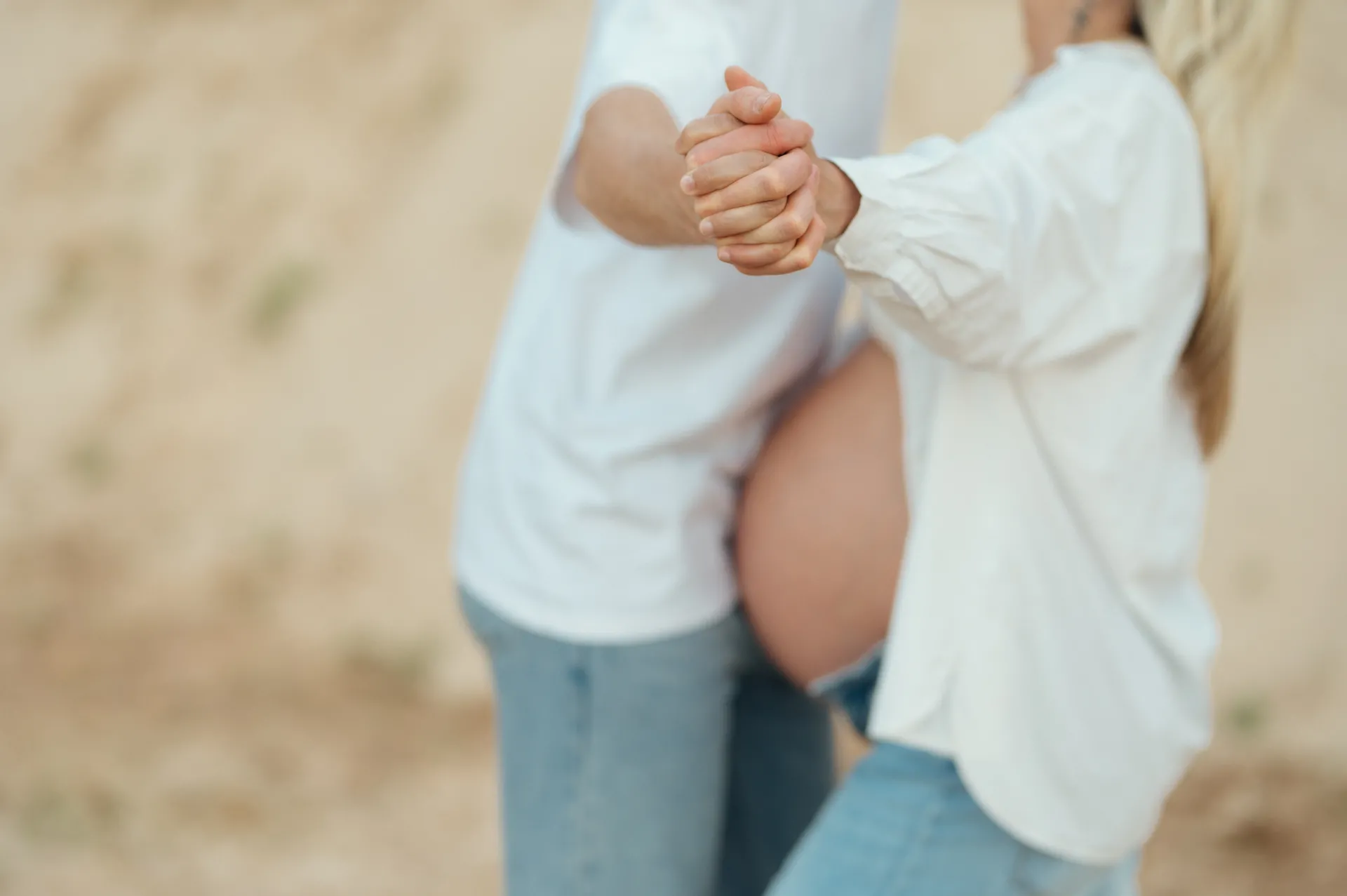 Grūtniece un viņas partneris, tērpušies baltās kreklu un džinsos, tur viens otru aiz rokas brīvā dabā uz maiga, smilšaina fona — maigs grūtnieces portrets.