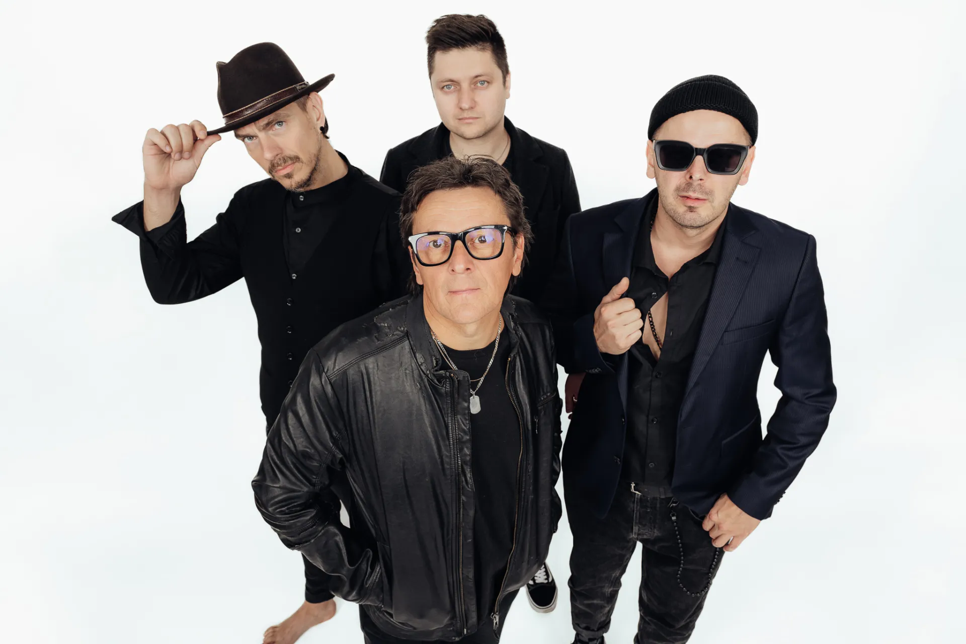 A high-angle studio portrait of four men posing together against a plain white background. The man in the foreground wears a black leather jacket and glasses, while the others behind him wear dark clothing, a fedora hat, sunglasses and a beanie, creating a stylish, moody group portrait with even lighting.