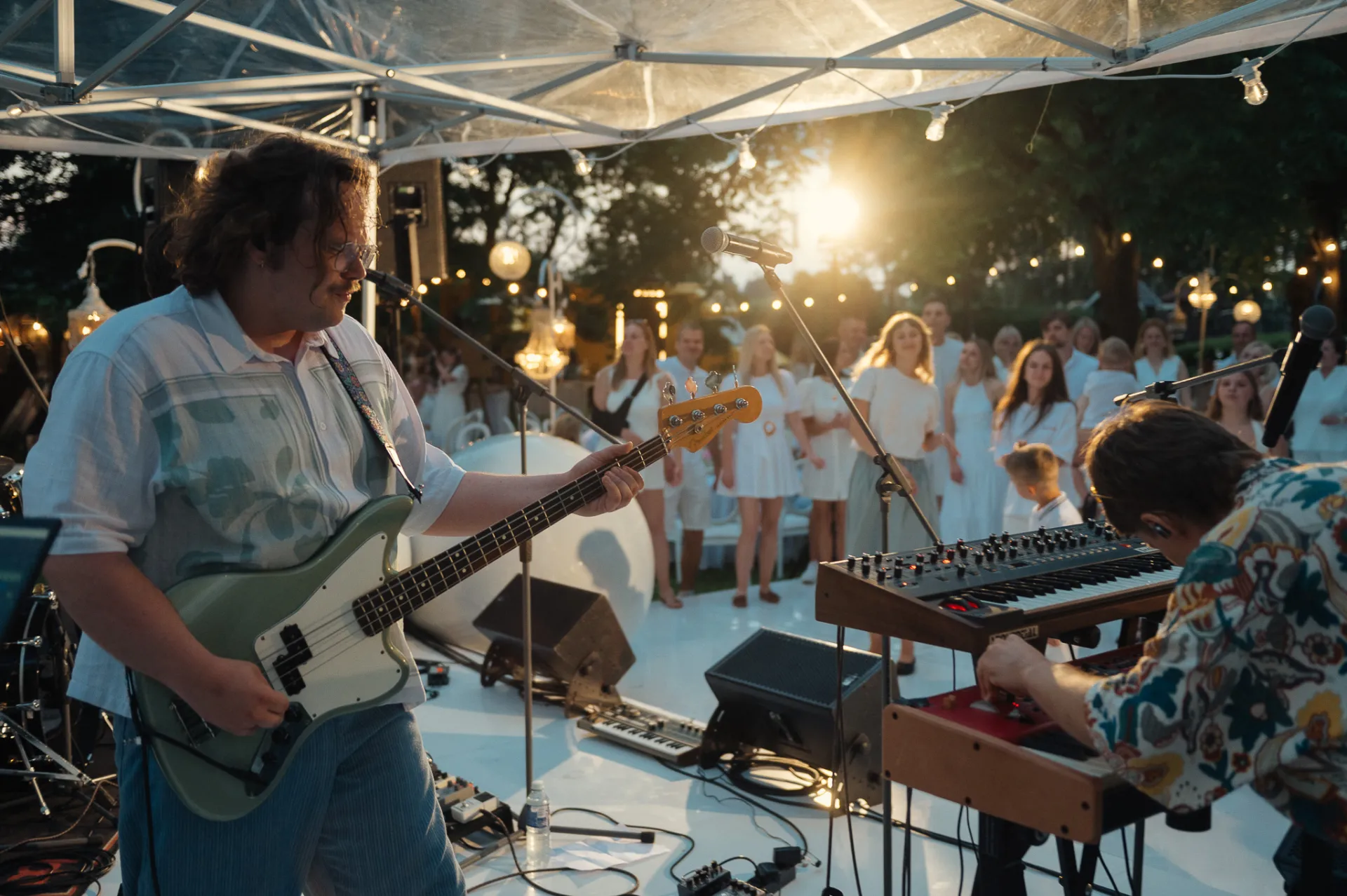 A band performs outdoors under a tent at dusk, with a guitarist in a patterned shirt playing a light green bass guitar in the foreground and a keyboardist at a synthesizer beside him, while a choir in white stands behind them against a warm sunset glow.