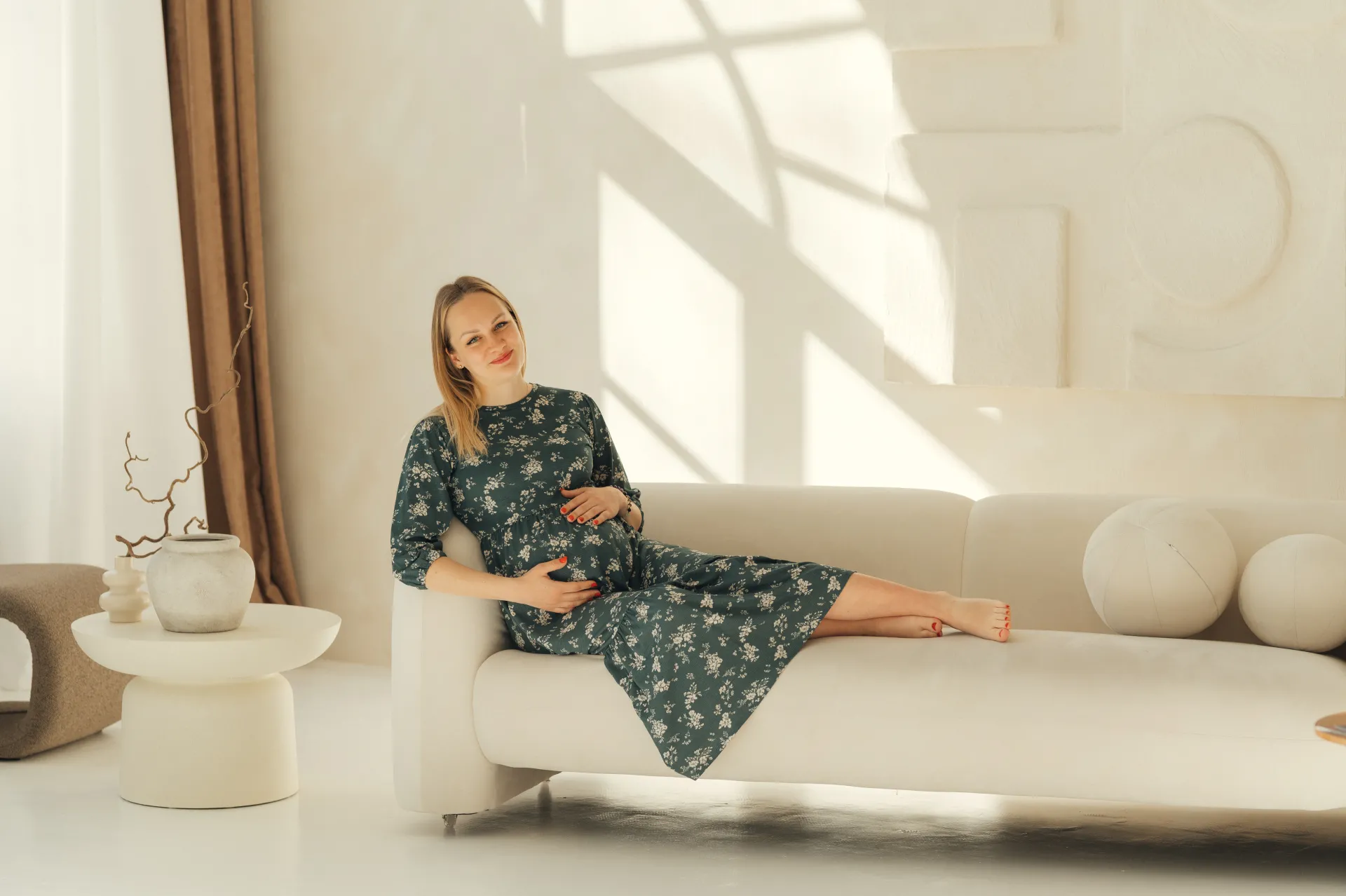 A pregnant woman in a dark green floral dress sits relaxed on a white sofa, hands resting on her belly, barefoot with legs extended. The bright minimal interior with soft natural light and patterned shadows creates a calm maternity portrait lifestyle scene.