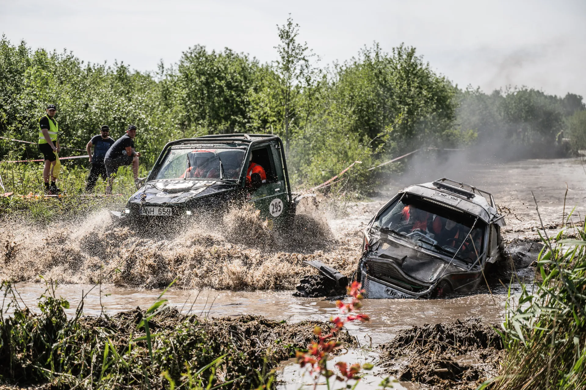 Divi apvidus automobiļi traucas cauri dubļainam ūdens šķērslim, izšļakstot brūnu ūdeni, kamēr skatītāji to vēro no zālāja krasta zem apmākušām debesīm.
