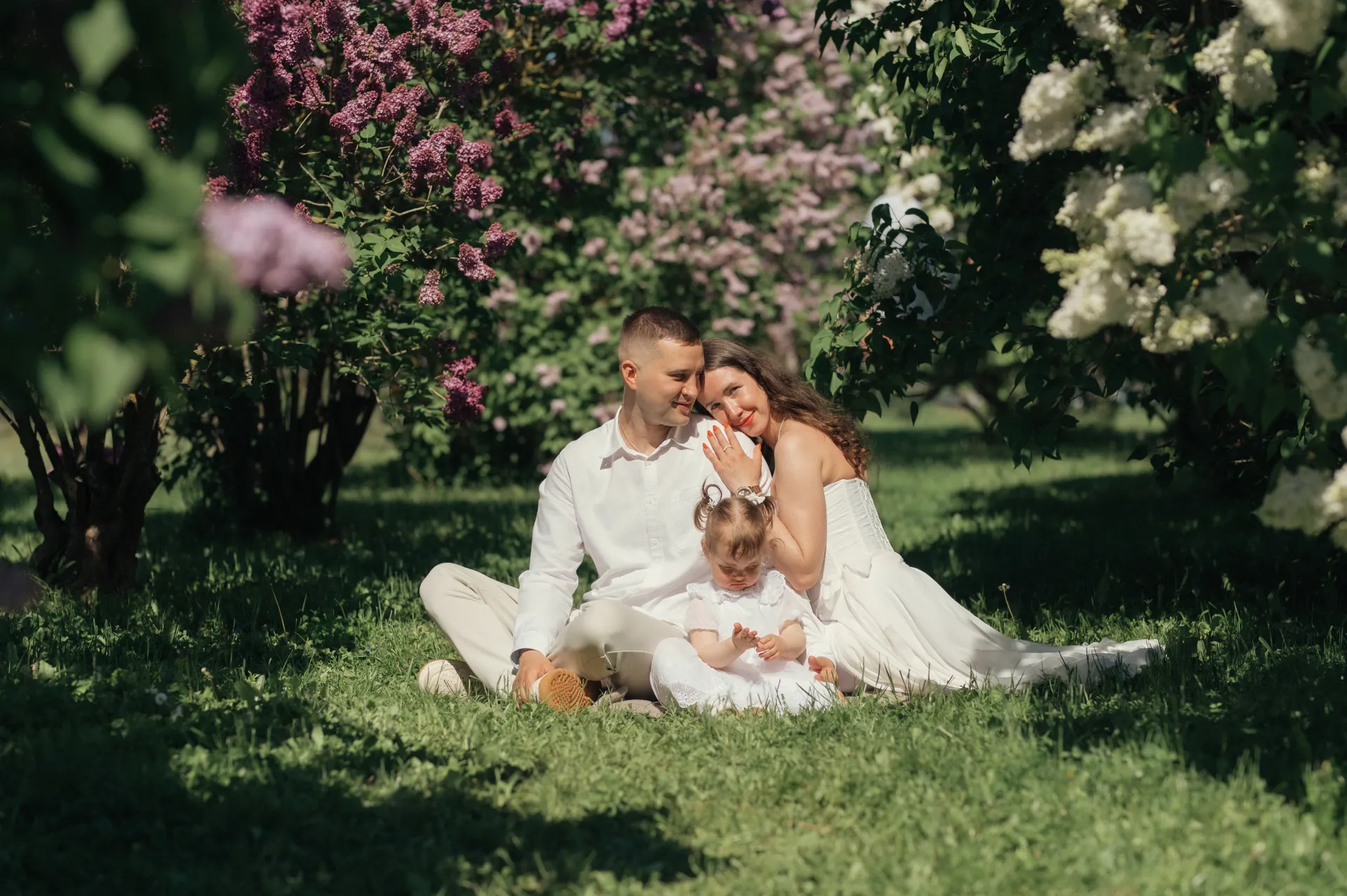 Ģimenes portretā redzams vīrietis, sieviete un mazs bērns, kas kopā sēž zālē zem ziedošiem ceriņu krūmiem, tērpušies baltā apģērbā un bauda sirsnīgu brīdi maigā, plankumainā saules gaismā.