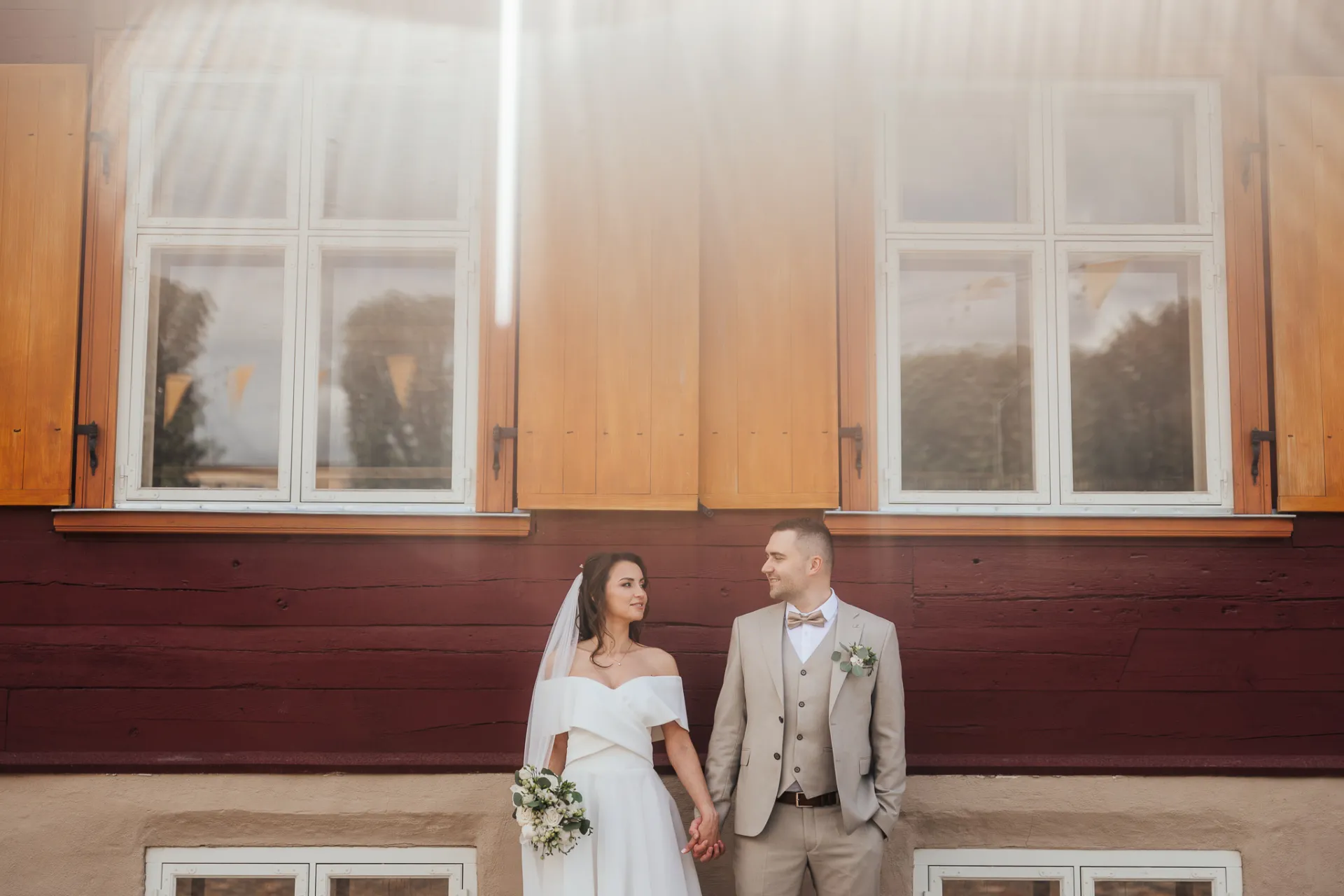 A bride in an off-the-shoulder white gown and veil holds hands with a groom in a light three-piece suit and bow tie, standing before a wooden building with large windows and bright sun flare.