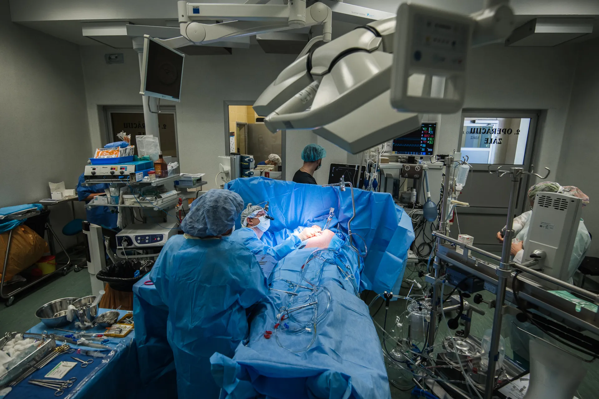 Ķirurģiskā komanda veic operāciju spilgti apgaismotā operāciju zālē, kur medicīnas personāls, tērpies zilos operācijas tērpos un cepurēs, apstājas ap pacientu, kurš ir pārklāts ar steriliem pārklājiem zem operāciju zāles apgaismojuma.