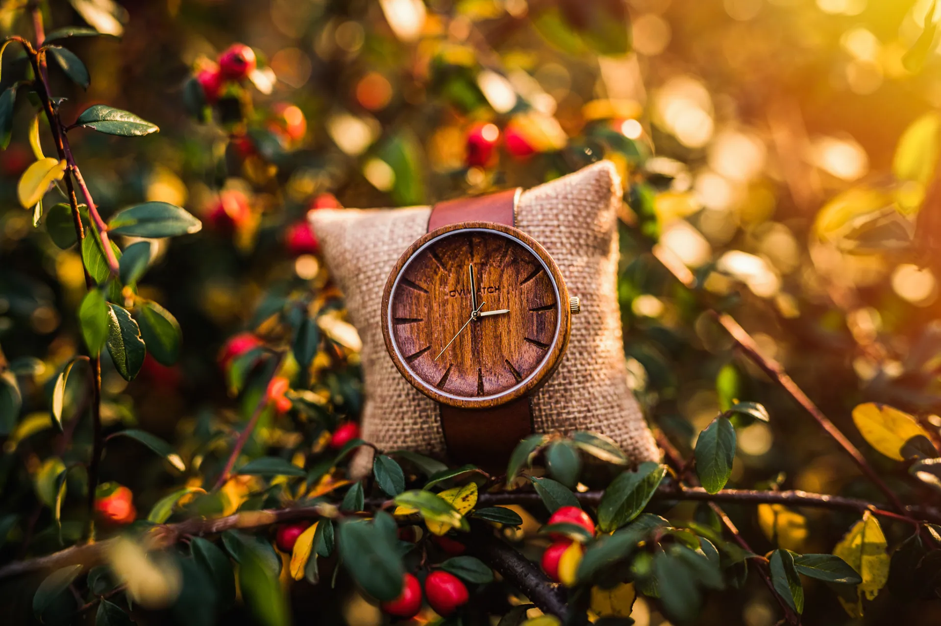 A wooden wristwatch with a brown leather strap rests on a burlap cushion amid green leaves and red berries, bathed in warm golden sunlight with soft bokeh in the background.