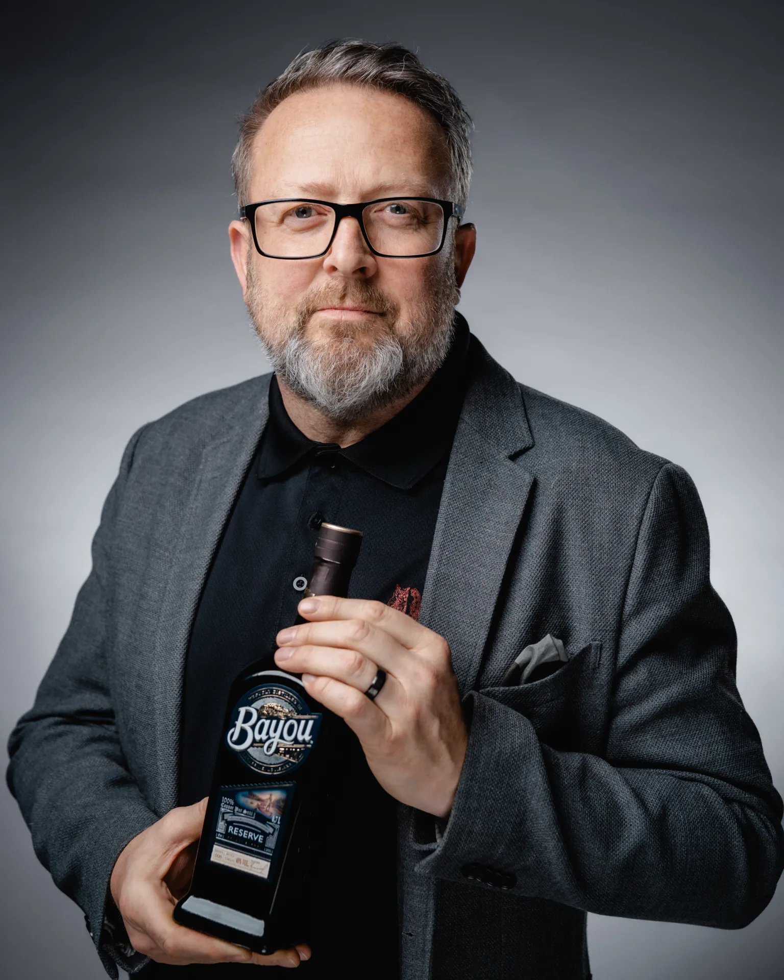 Biznesa Portreti — A middle-aged man with glasses and a gray beard wears a gray blazer over a black polo and holds a dark bottle with a visible label in front of him. The studio portrait uses soft gradient gray lighting for a clean commercial look with a neutral, professional mood.