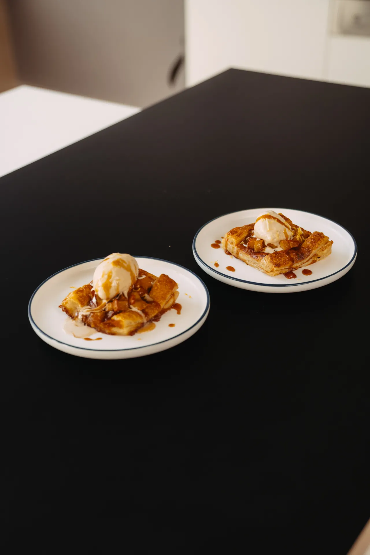 Ēdiena Fotogrāfija — Two white plates with blue rims sit on a black tabletop, each holding a golden-brown puff pastry topped with a scoop of vanilla ice cream and drizzled caramel sauce. The minimal interior setting and soft, warm lighting create a calm, appetizing food still life in a commercial s…
