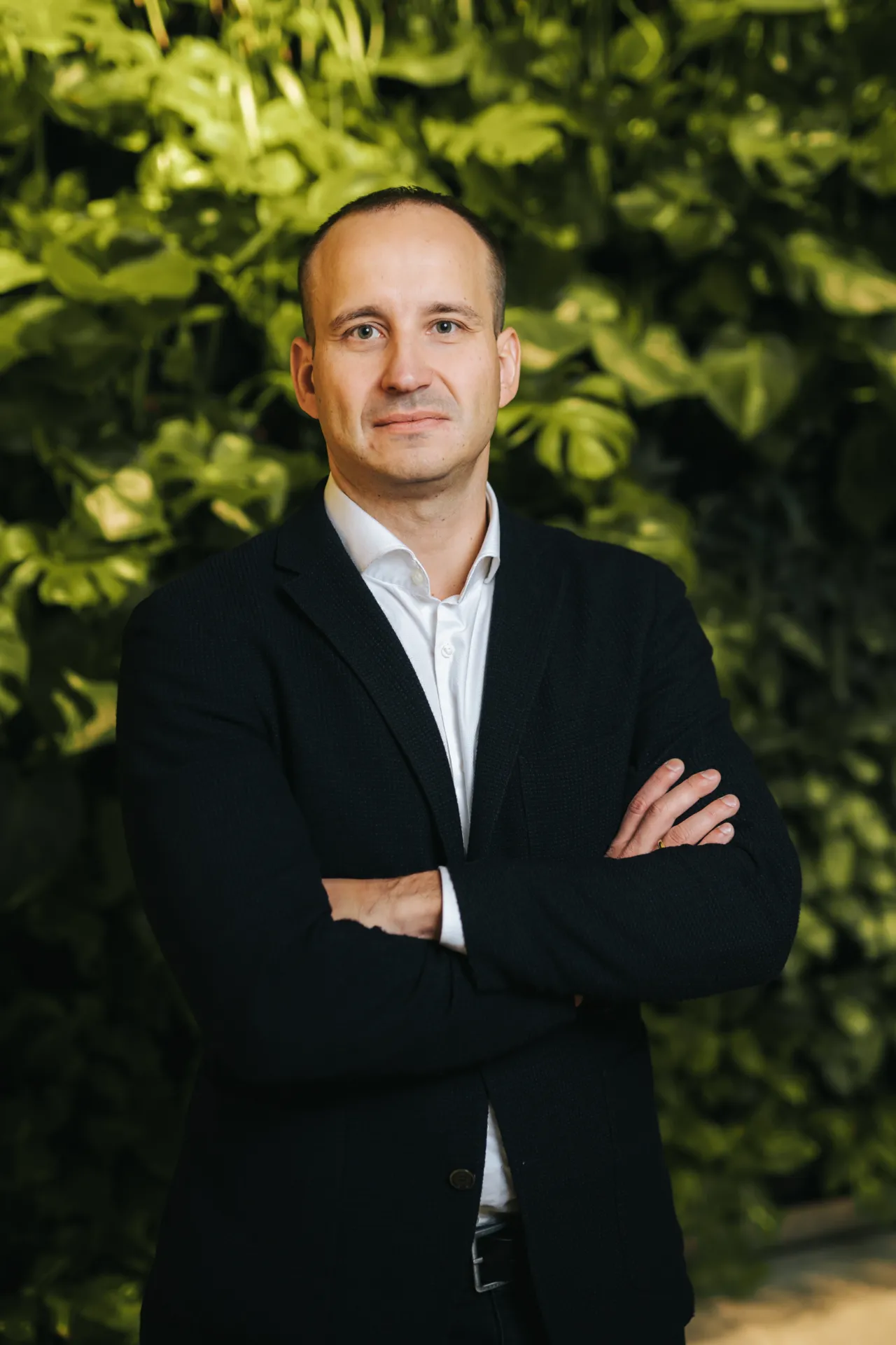 Biznesa Portreti — A middle-aged man with short hair stands facing the camera with his arms crossed, wearing a dark blazer over a white collared shirt. He is positioned in front of a dense green living wall backdrop, lit with soft natural light in a formal corporate portrait style.
