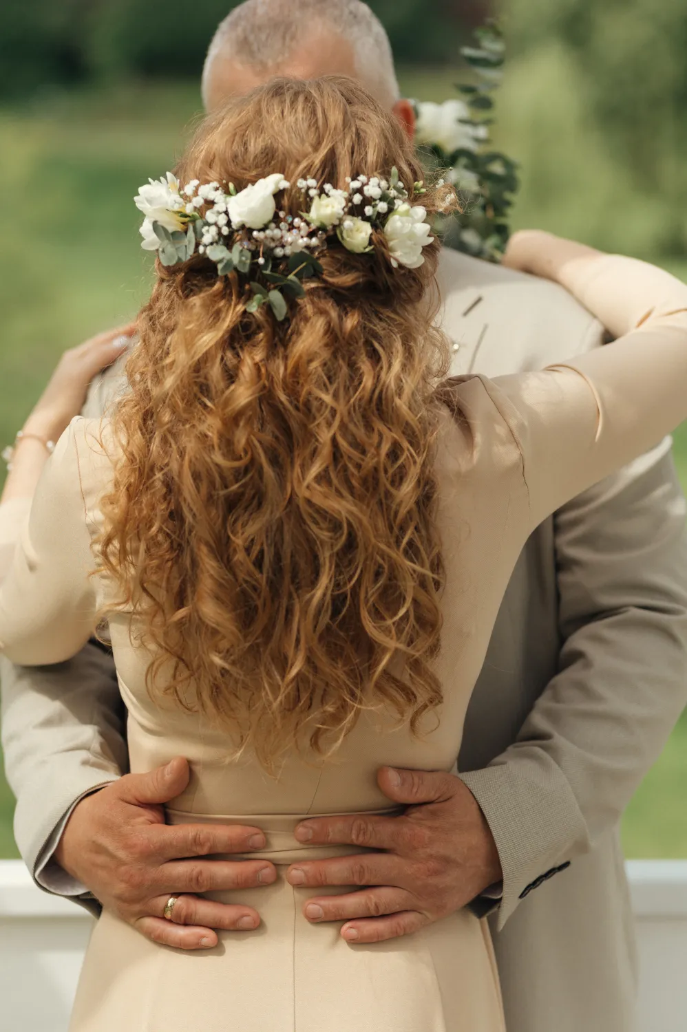 A bride with long curly hair and a floral crown embraces a groom in a light suit, their hands resting on her waist against a soft green outdoor background.