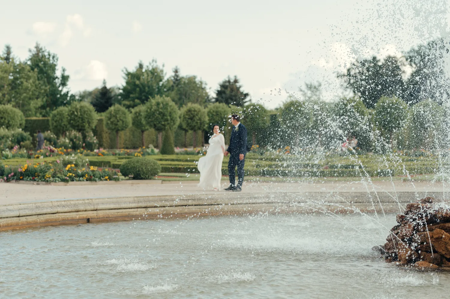 A bride in a white dress and a groom in a dark suit walk hand in hand across a garden path in the background, with a fountain spraying water in the foreground. Soft daylight illuminates manicured hedges, flowerbeds and trees, giving the scene a romantic wedding portrait mood.