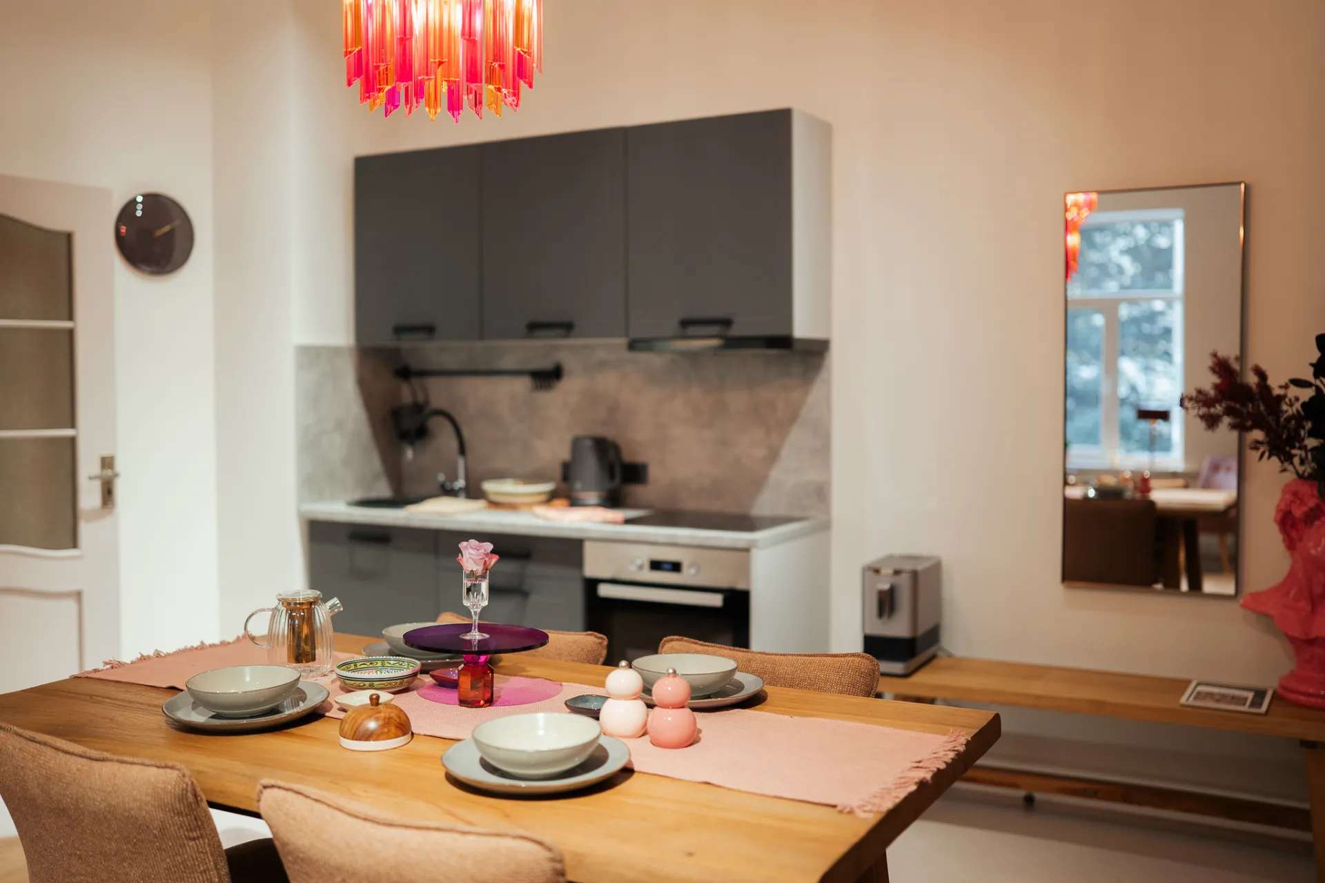 A warmly lit modern kitchen and dining area with a wooden table set for dining, pink runner, bowls and plates, and a colorful pendant light overhead. Dark gray cabinets, a built-in oven, mirror on the wall, and a wooden bench create a cozy lifestyle interior scene, captured in a real estate-style photograph.
