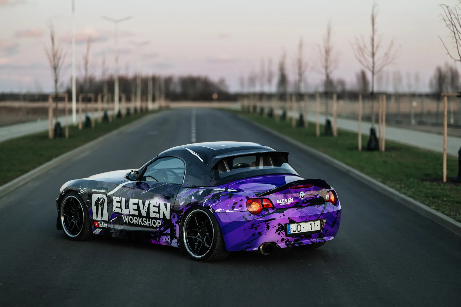 A rear three-quarter view of a purple and black wrapped sports convertible with a rear spoiler and custom wheels parked on an empty asphalt road at dusk. Young trees line the grassy verge beside the road under a soft pinkish sky, giving a calm automotive editorial style.