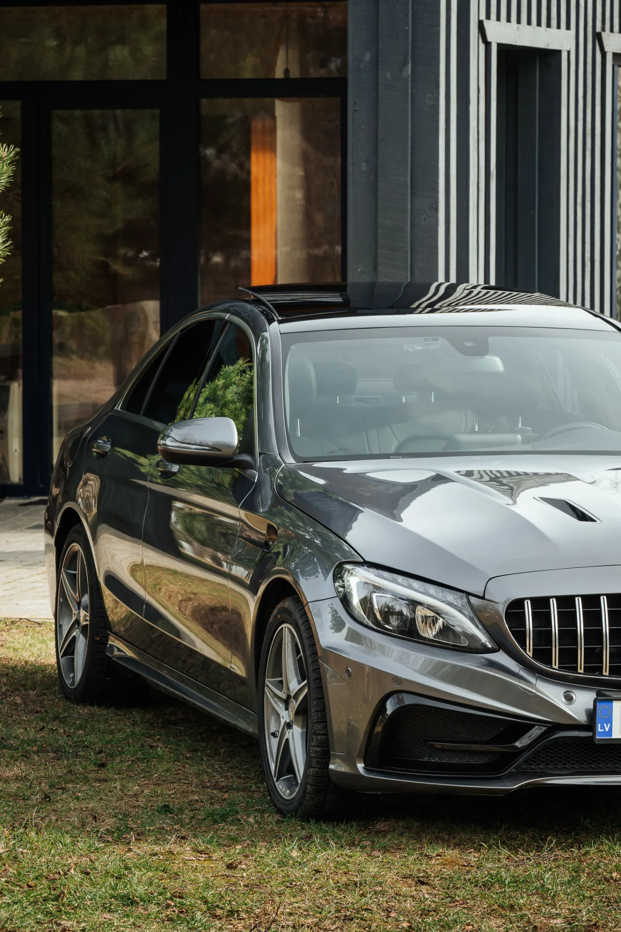 A silver sedan is parked on grass in front of a modern building with dark vertical siding and large glass windows. The car features a prominent front grille, alloy wheels, and a sunroof, captured in bright daylight in an automotive photography style.