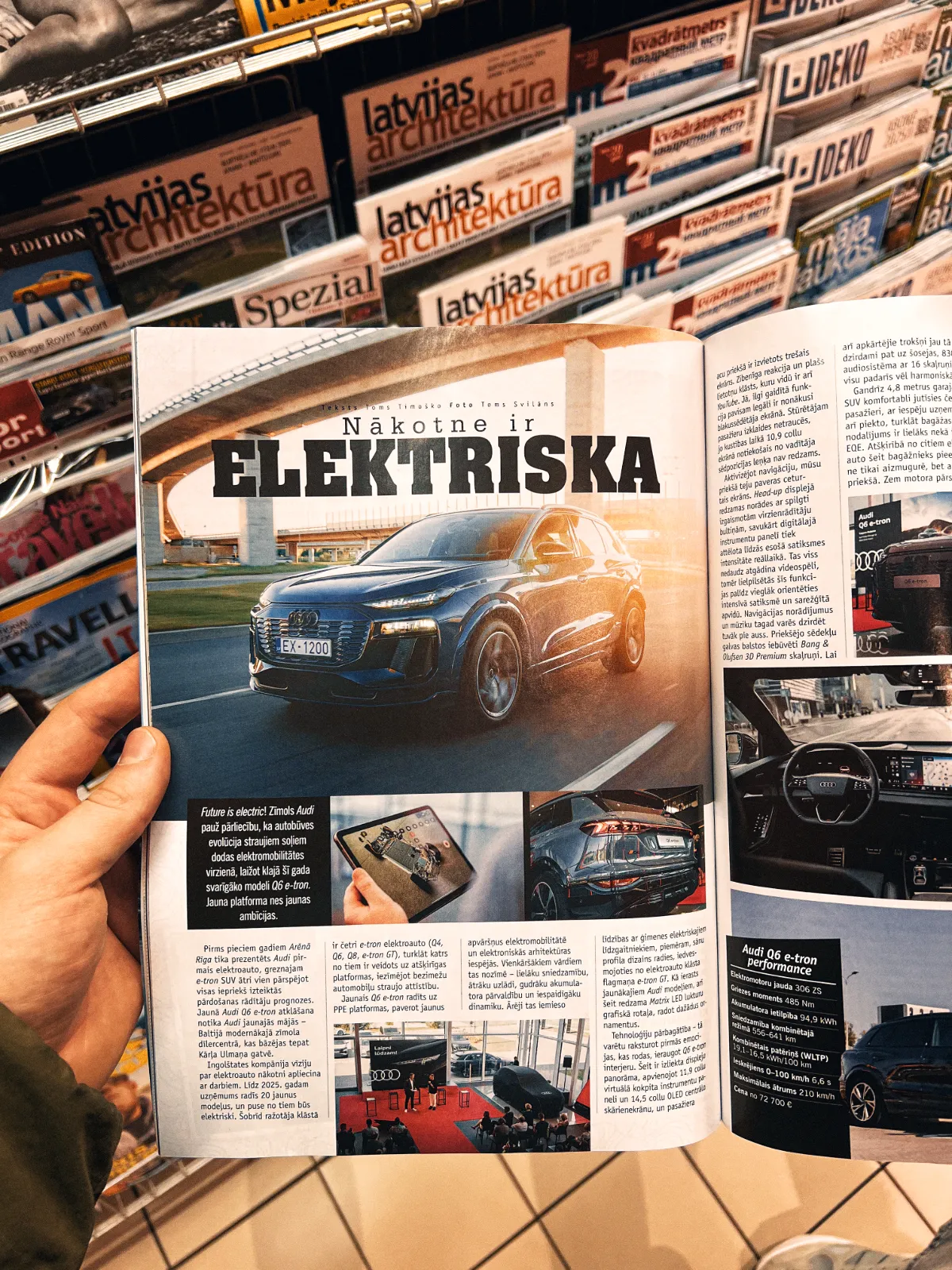 Auto Foto — A person’s hand holds an open magazine showing a glossy spread about an electric car with the large headline “Nākotne ir ELEKTRISKA” and smaller images below. Behind it, shelves are lined with stacked magazines; the scene is a casual editorial lifestyle shot with warm natural light and …