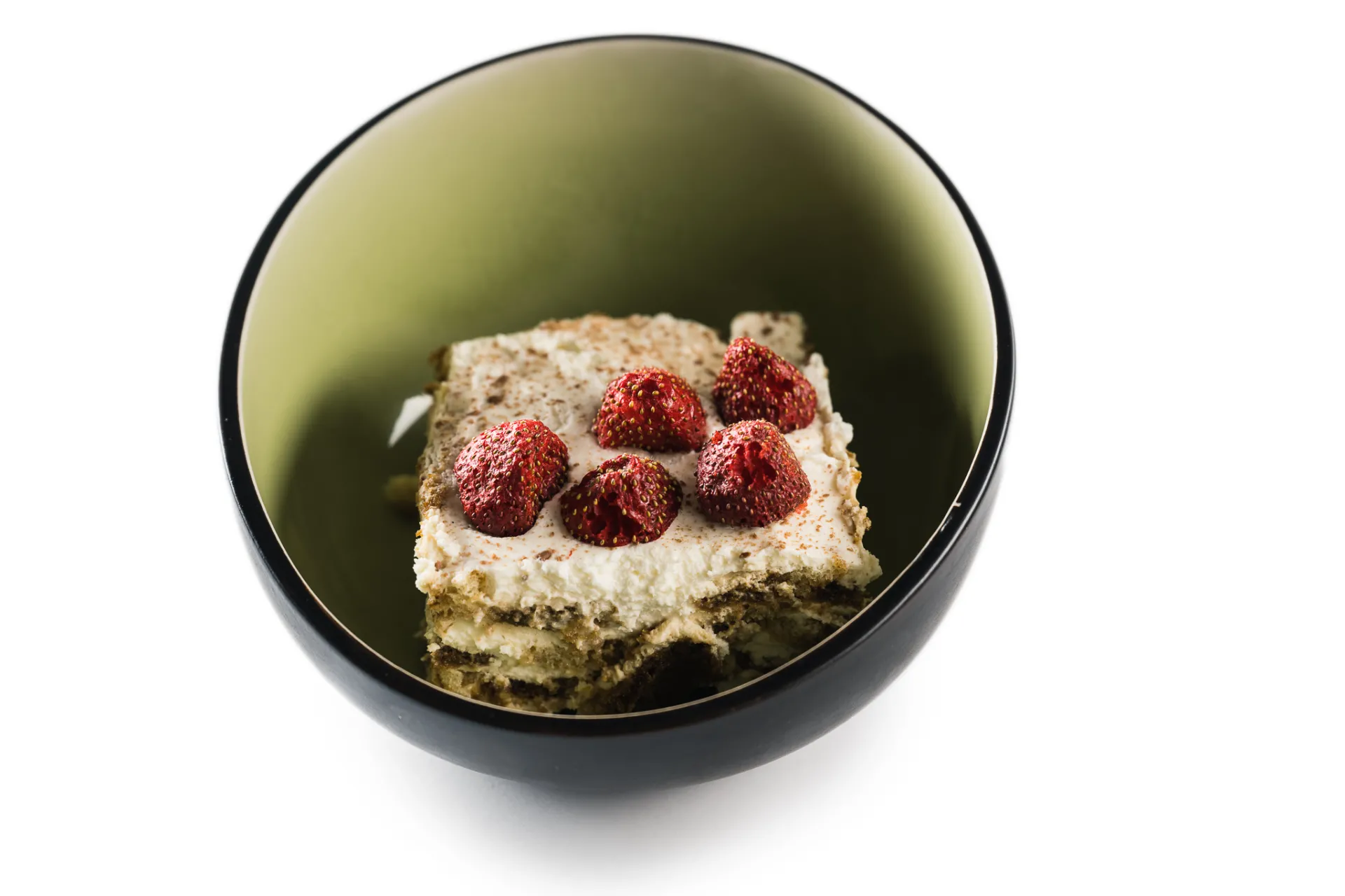 Kvadrātveida gabals slāņveida tiramisu, kas rotāts ar svaigām zemenēm un kakao pulveri, atrodas bļodā ar tumšu malu un olīvzaļu iekšpusi; tas ir nofotografēts uz tīra balta fona komerciālā pārtikas fotogrāfijas stilā.