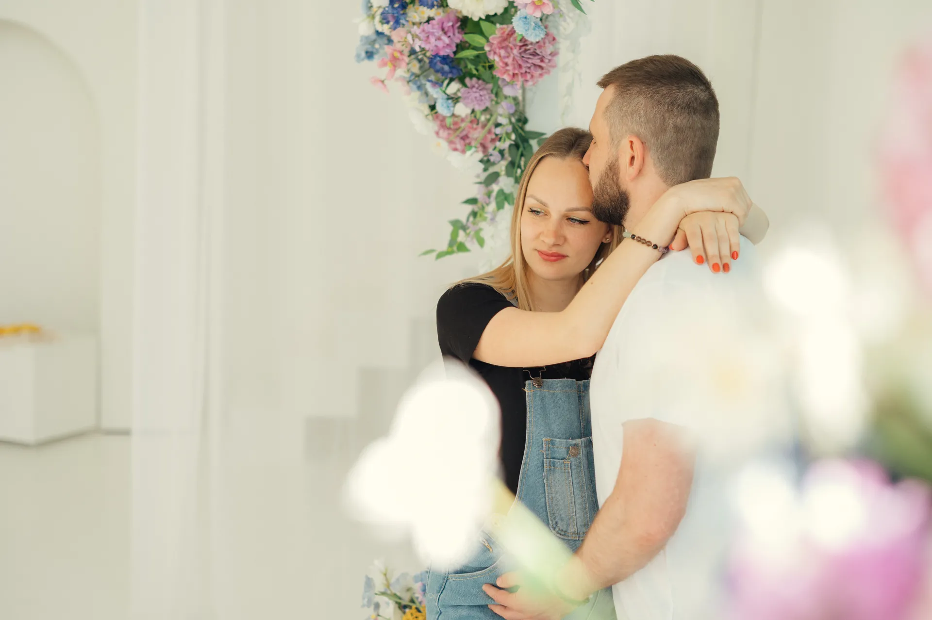Pāris sirsnīgi apskauj viens otru gaišā, minimālisma stilā iekārtotā baltā studijā, ko rotā pasteļtoņu ziedu kompozīcijas. Sieviete melnā topā un džinsu kombinezona apģērbā atbalsta galvu pret vīrieti baltā T-kreklā, un maigā dabiskā gaisma rada siltu, romantisku portreta noskaņu.