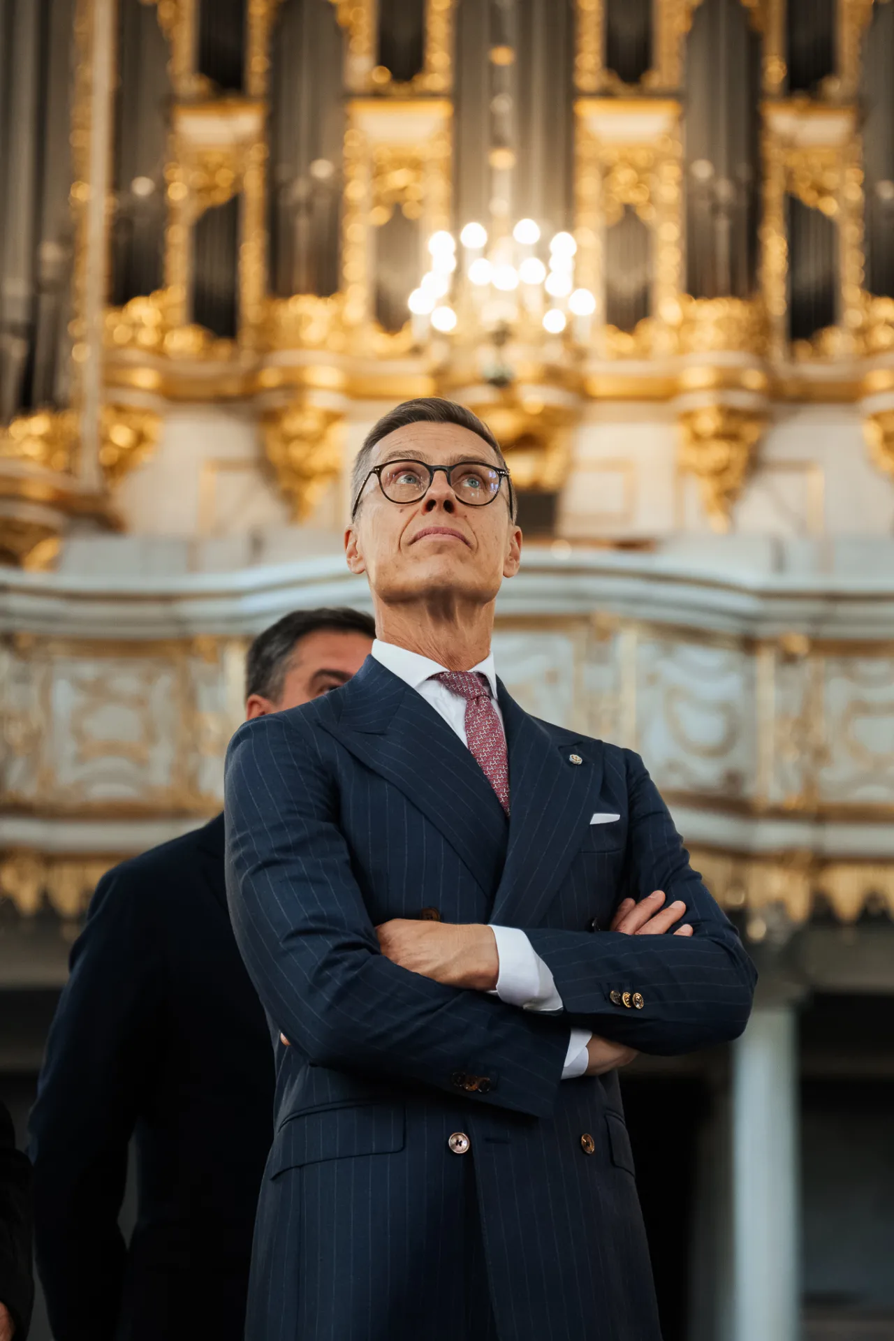 Vīrietis tumši zilā svītrainā uzvalkā un brillēs stāv ar sakrustotām rokām, skatīdamies uz augšu greznā zālē, kuras fonā redzamas zelta ērģeles un lustra; formāls portreta stils.