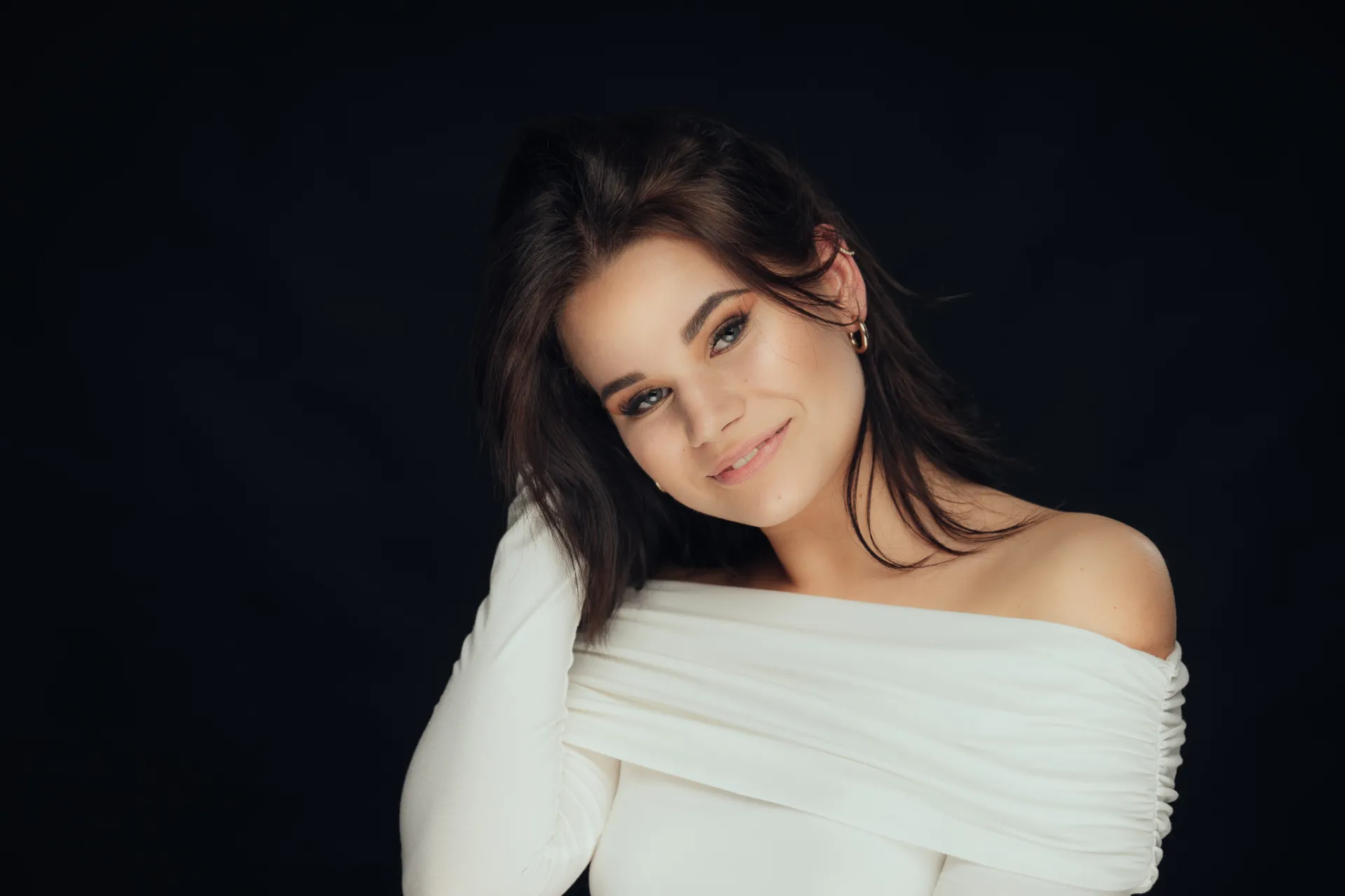 A young woman with long dark brown hair poses against a solid black background, wearing a white off-the-shoulder long-sleeve top. She is turned slightly toward the camera with a soft smile, her head tilted and hand raised near her hair in a studio portrait with dramatic lighting and a moody, elegant mood.