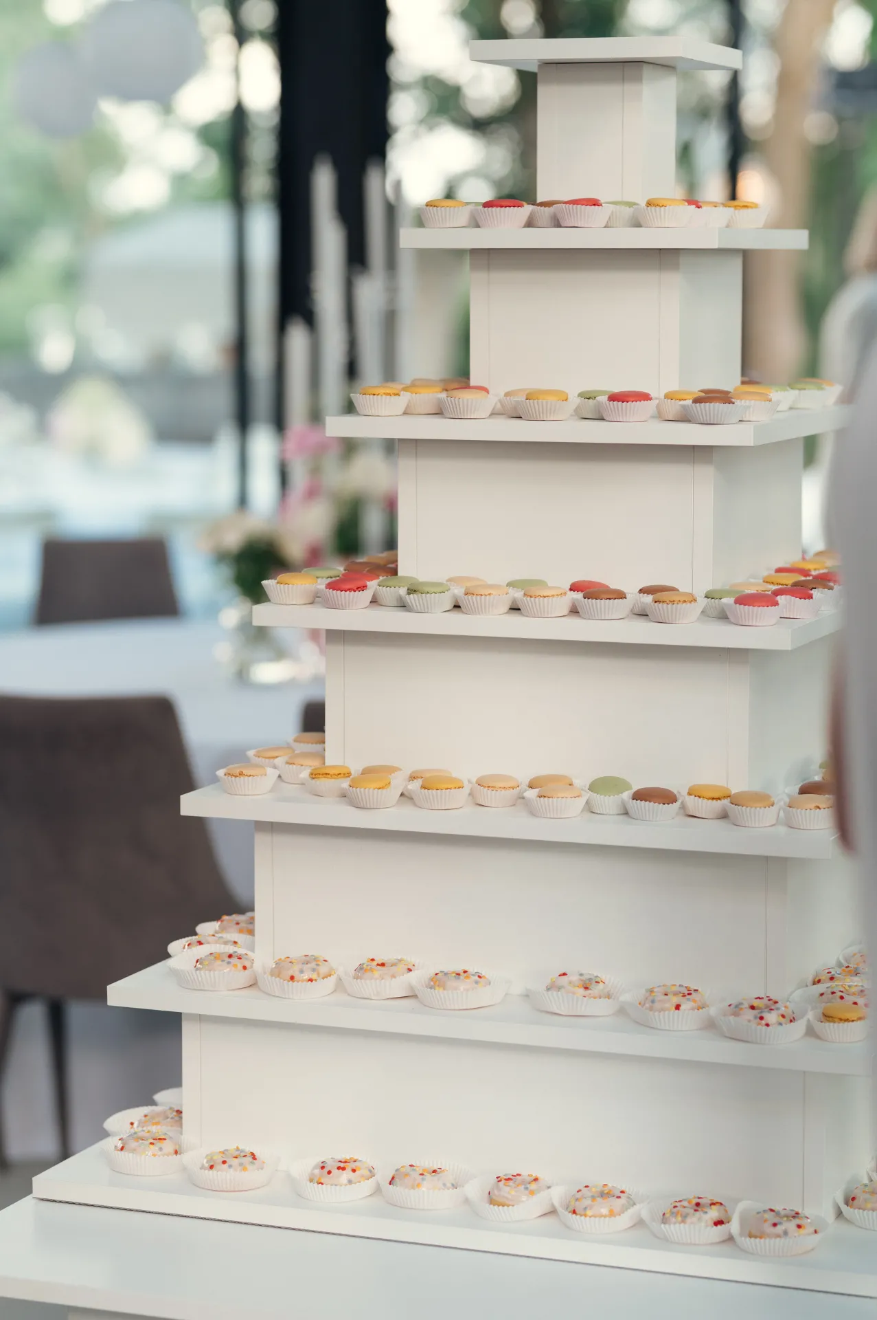 A white tiered dessert stand holds rows of colorful macarons and cupcakes with sprinkles, set indoors near a window with soft natural light and blurred greenery in the background.
