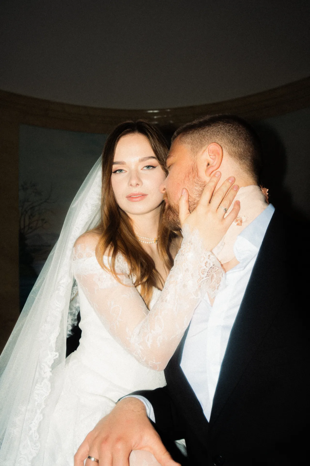 A bride in a white lace gown and veil looks at the camera while a groom in a black suit kisses her cheek, his hand resting on her arm in an indoor portrait setting.
